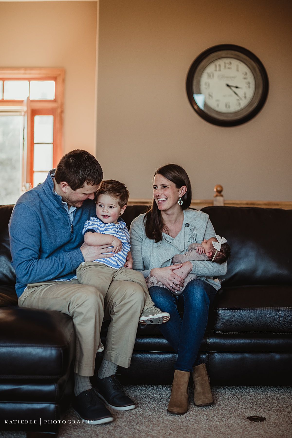 Family sitting on leather couch under wall clock during lifestyle photography session at home.