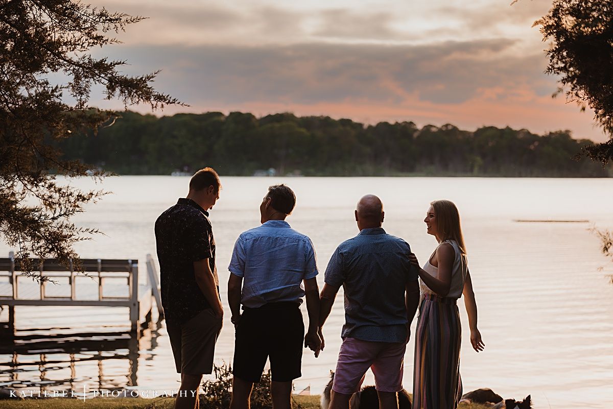 Silhouettes of four people standing on a dock at sunset overlooking a peaceful lake.