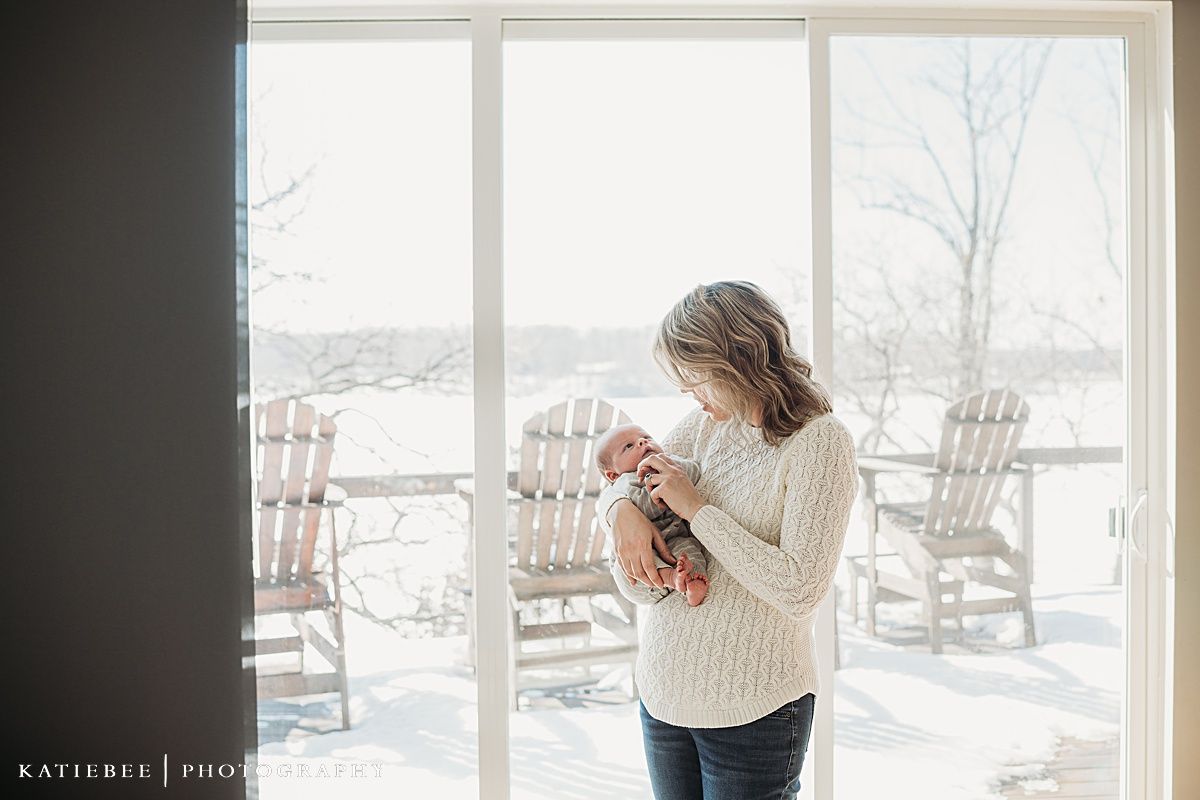 Parent in cable knit sweater holds baby while standing by windows with rocking chairs visible.