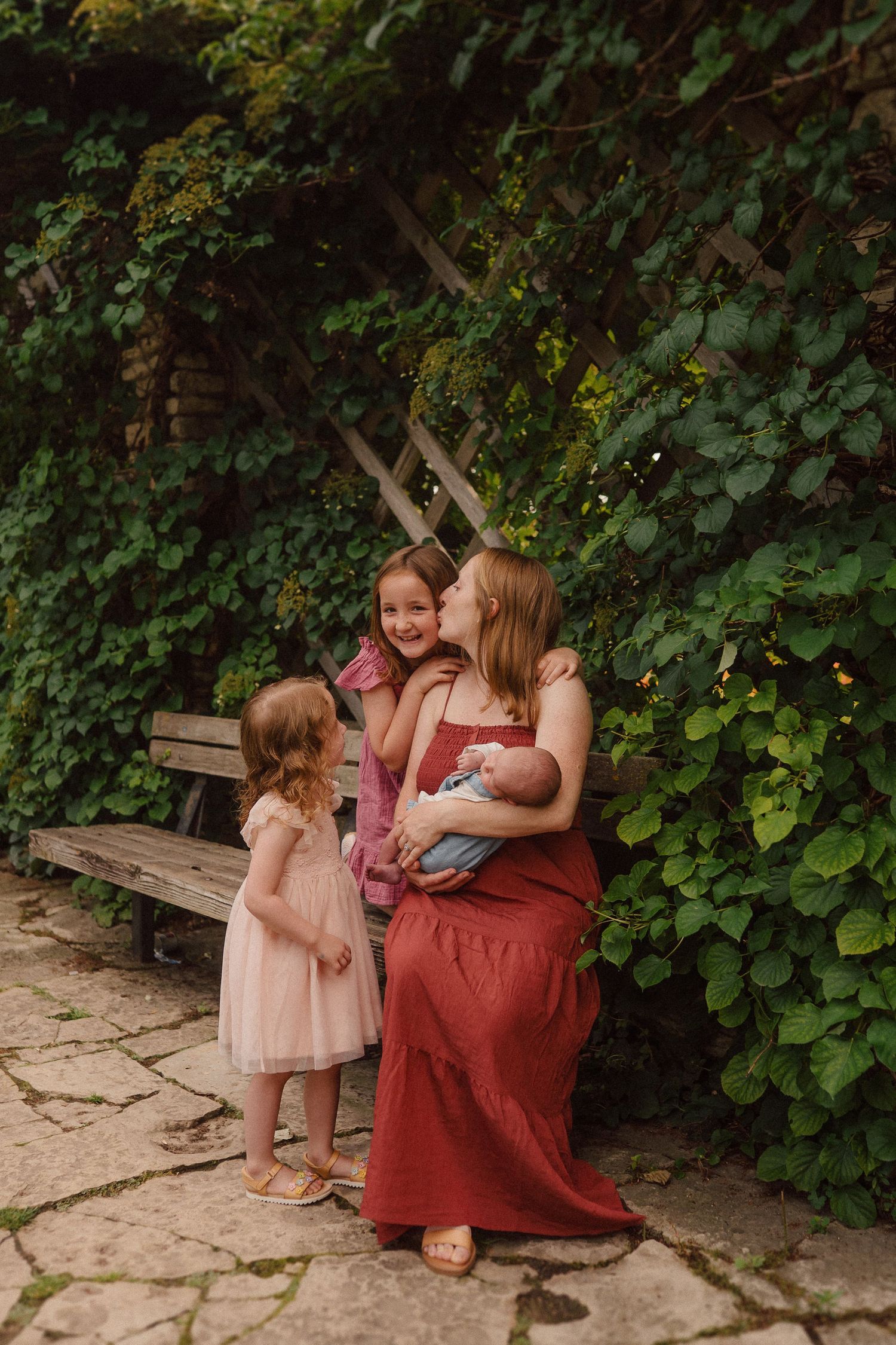 A family in coordinating red and pink outfits sits together on a wooden bench surrounded by lush green foliage.