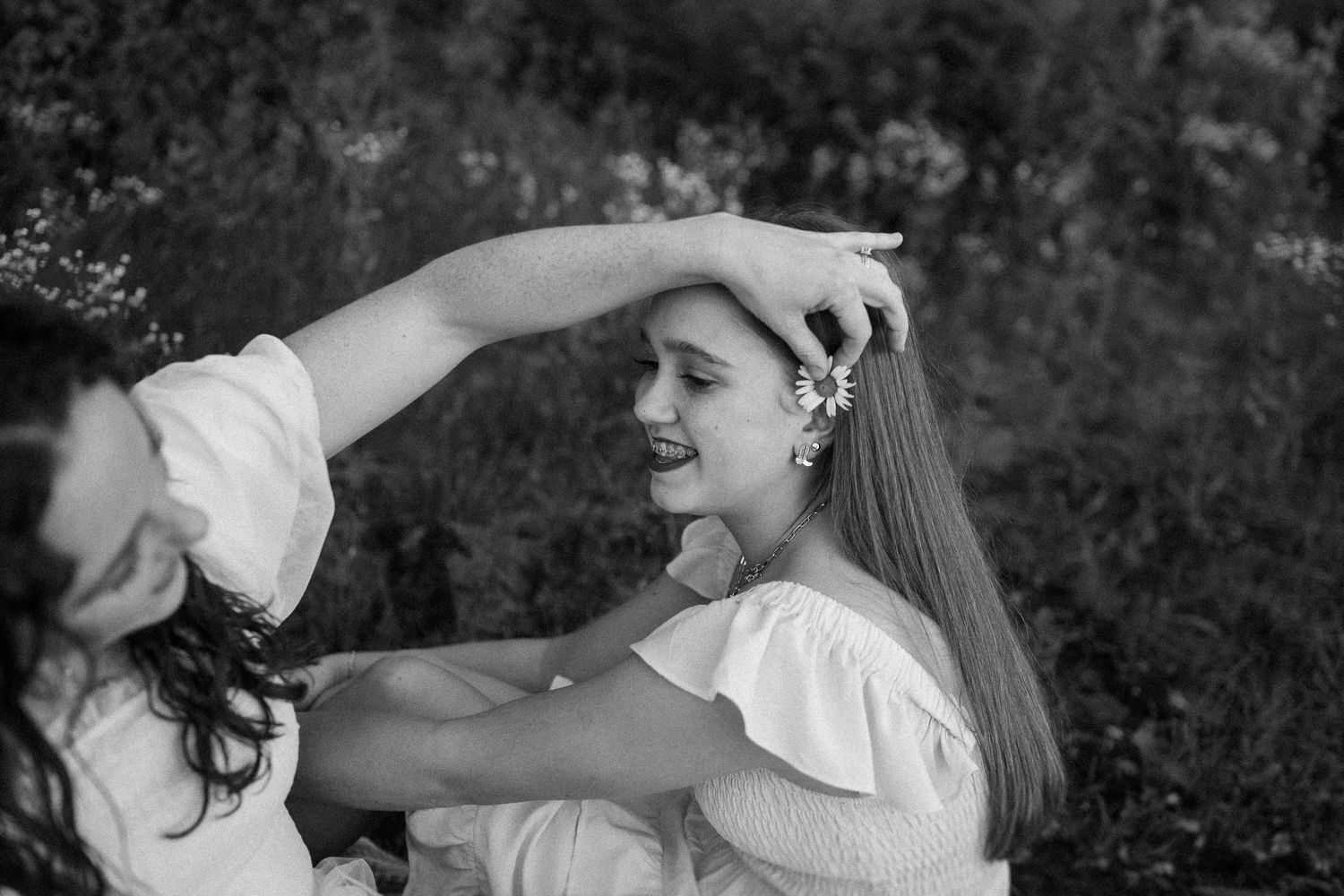An intimate black and white moment captured between friends wearing white dresses in a garden setting.