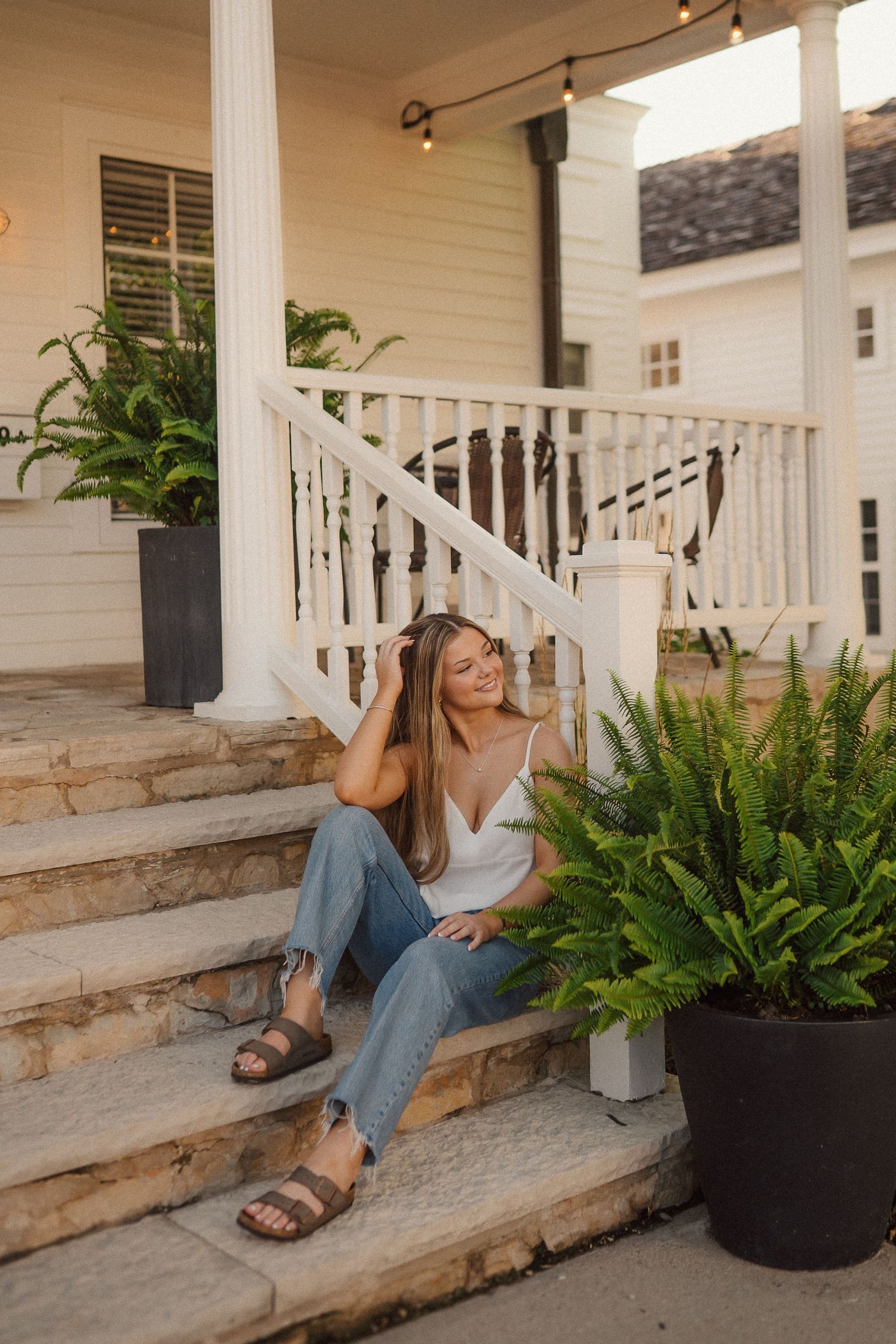 Person in casual attire sits on stone steps of a white porch surrounded by potted ferns.