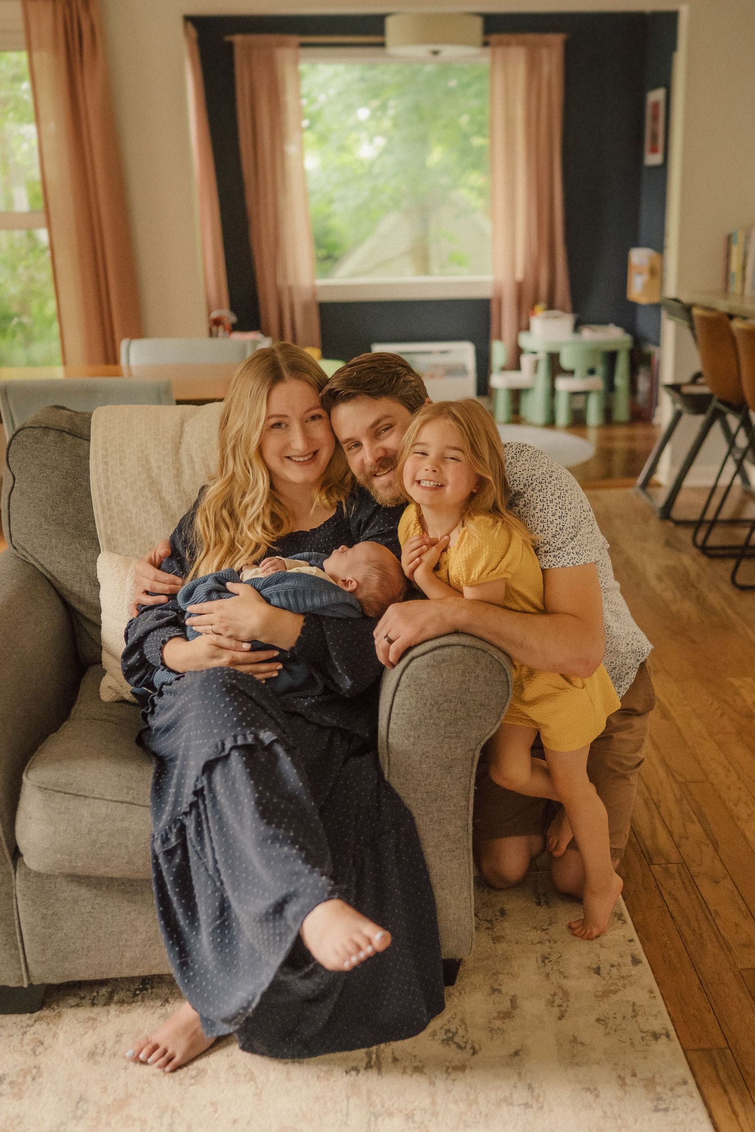 A family shares a joyful moment together on their grey couch while holding a newborn baby.