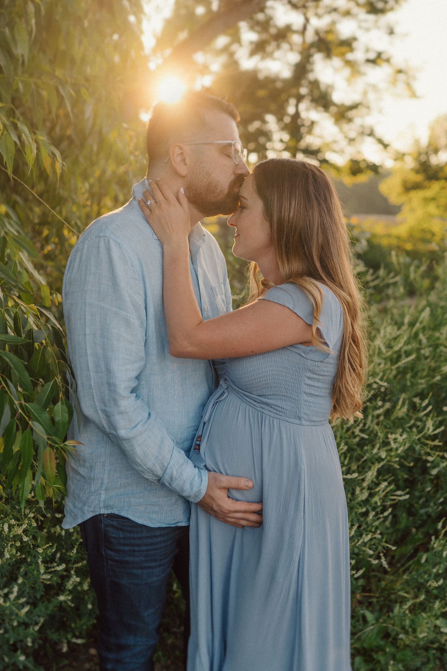 A romantic couple embraces in a light blue outfit against golden sunlight and lush greenery.