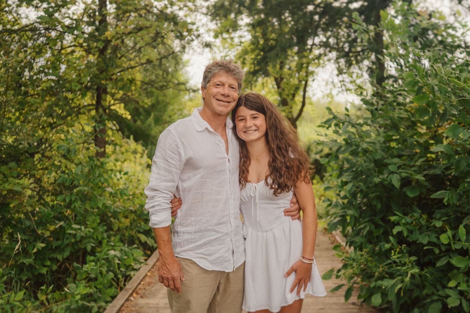 Couple in light colored outfits embrace while standing on rustic wooden path outdoors.