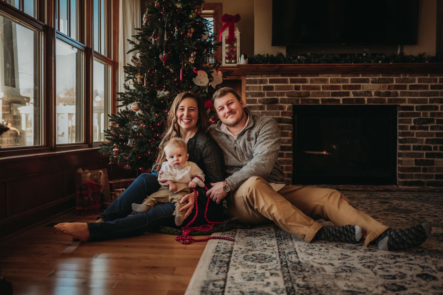 Family sitting together on floor near decorated Christmas tree and windows with natural light.