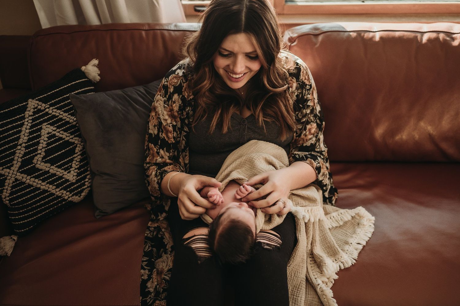 Mother in floral dress holding baby on brown leather couch with decorative pillows.
