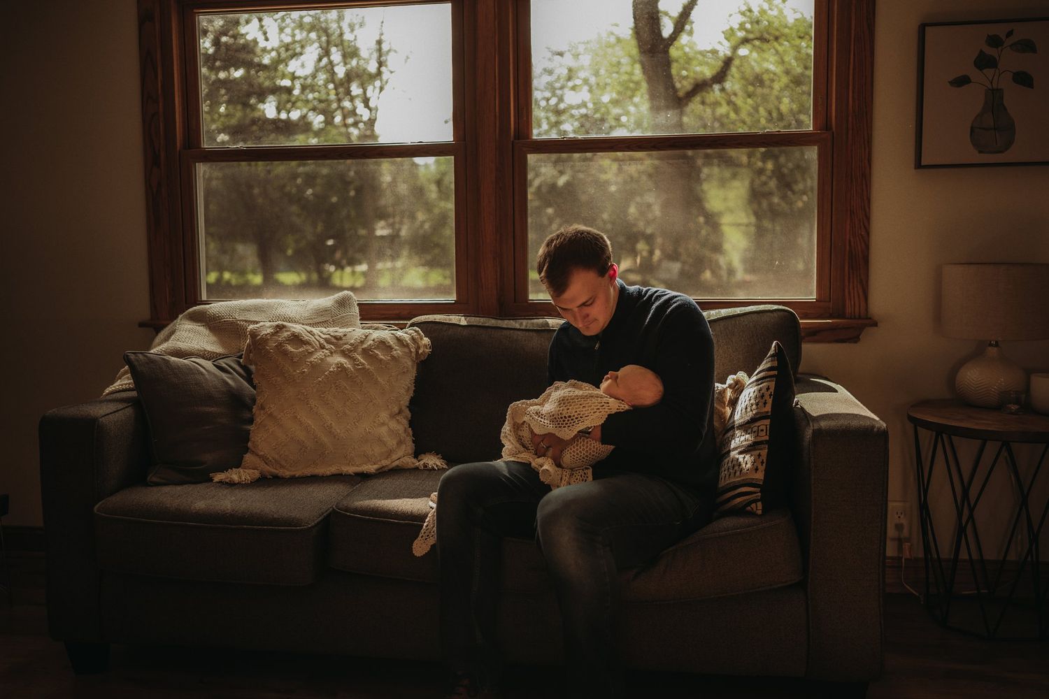 Someone sits on a couch by a window, cradling a baby in natural sunlight.