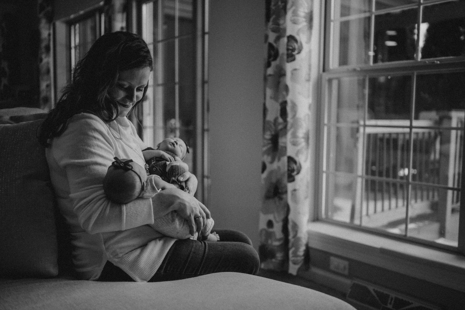 Black and white photo of parent with infant by window in serene living room setting.