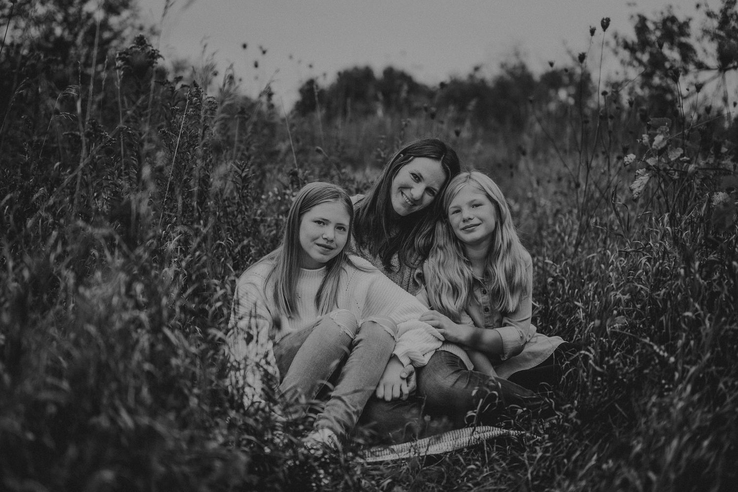 Black and white photo of three people sitting close together in wild grass field.