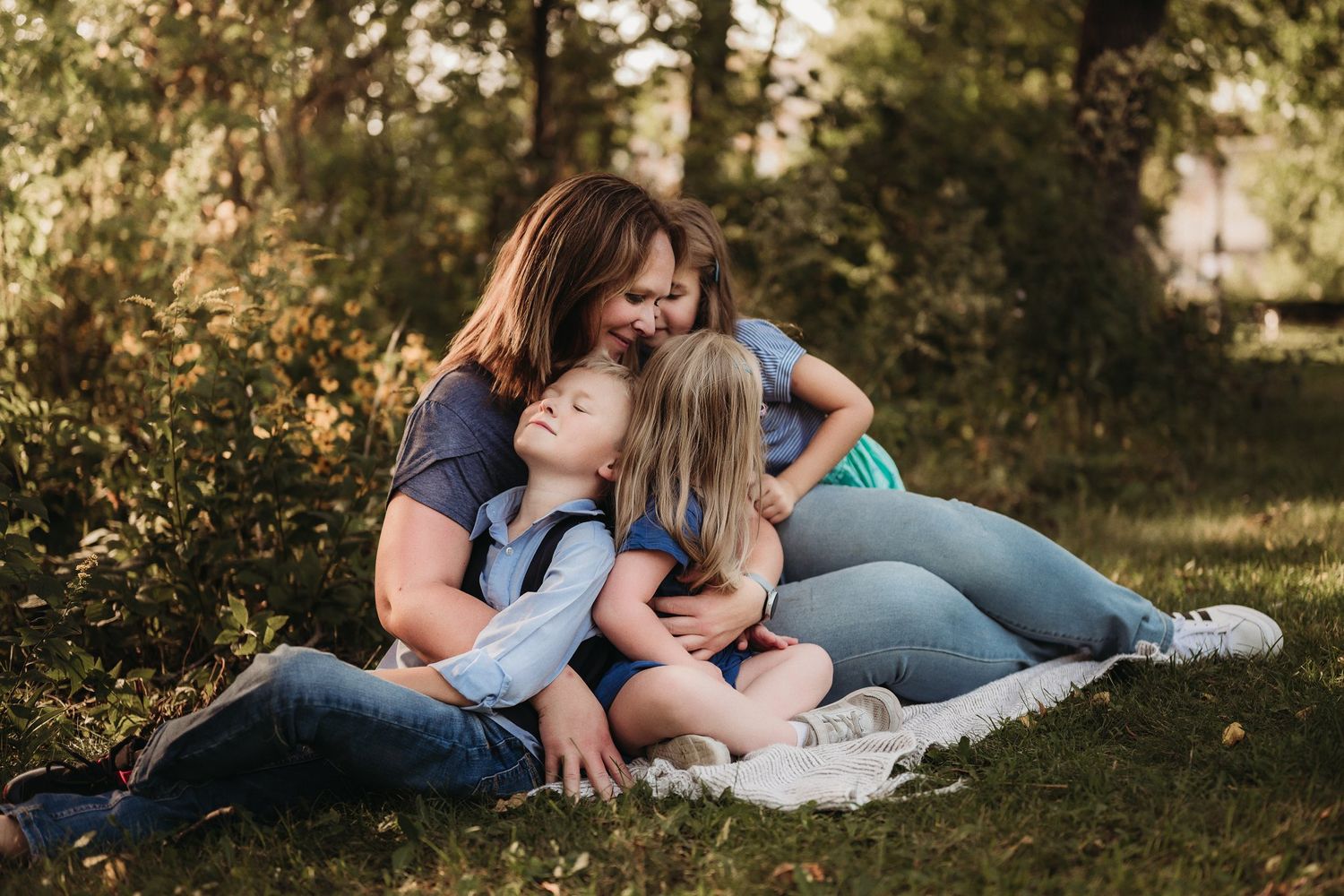 An intimate family moment shows loved ones embracing on a white blanket in dappled sunlight.