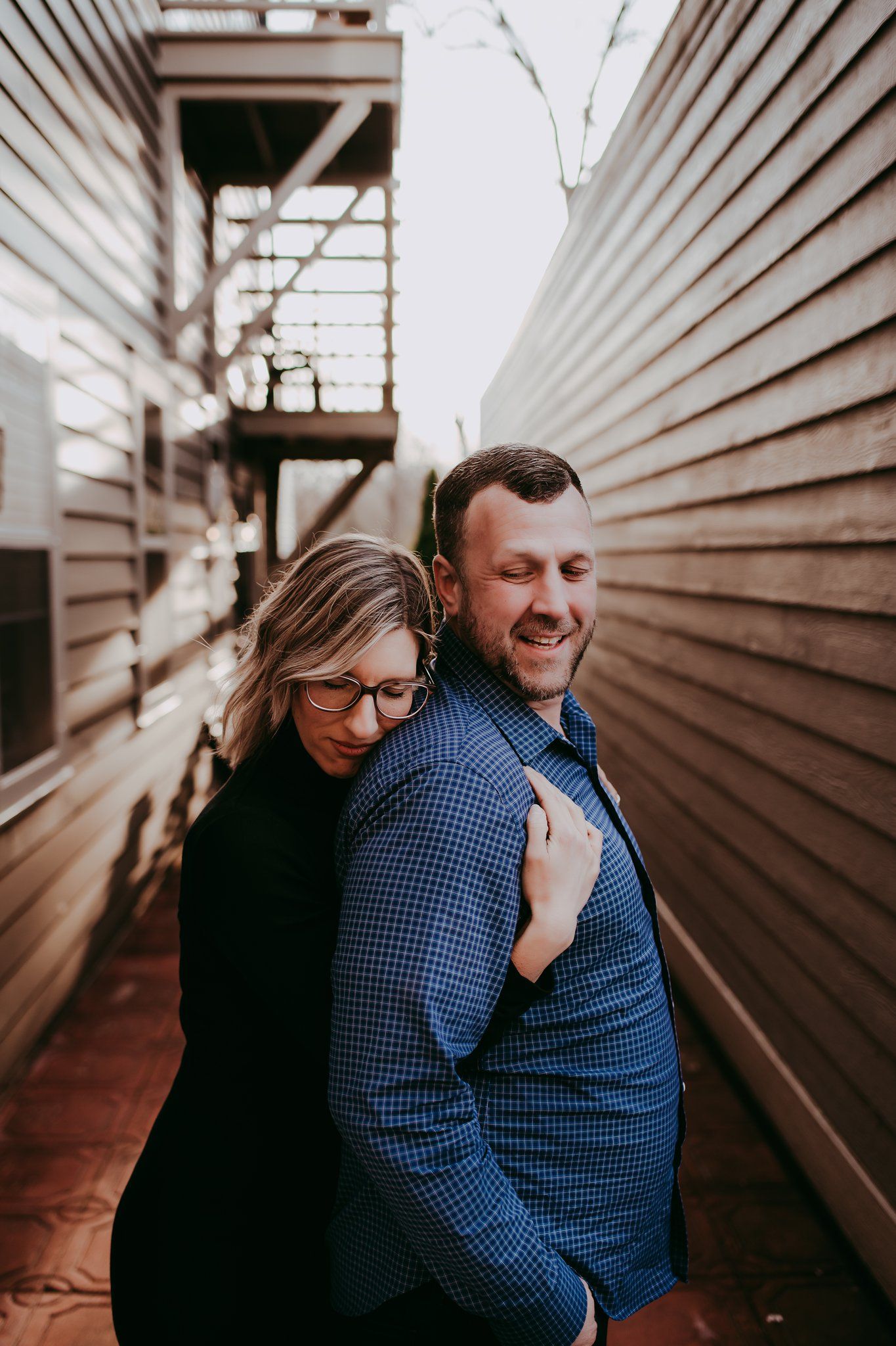 Romantic couple embrace in narrow walkway with wooden siding and natural lighting.