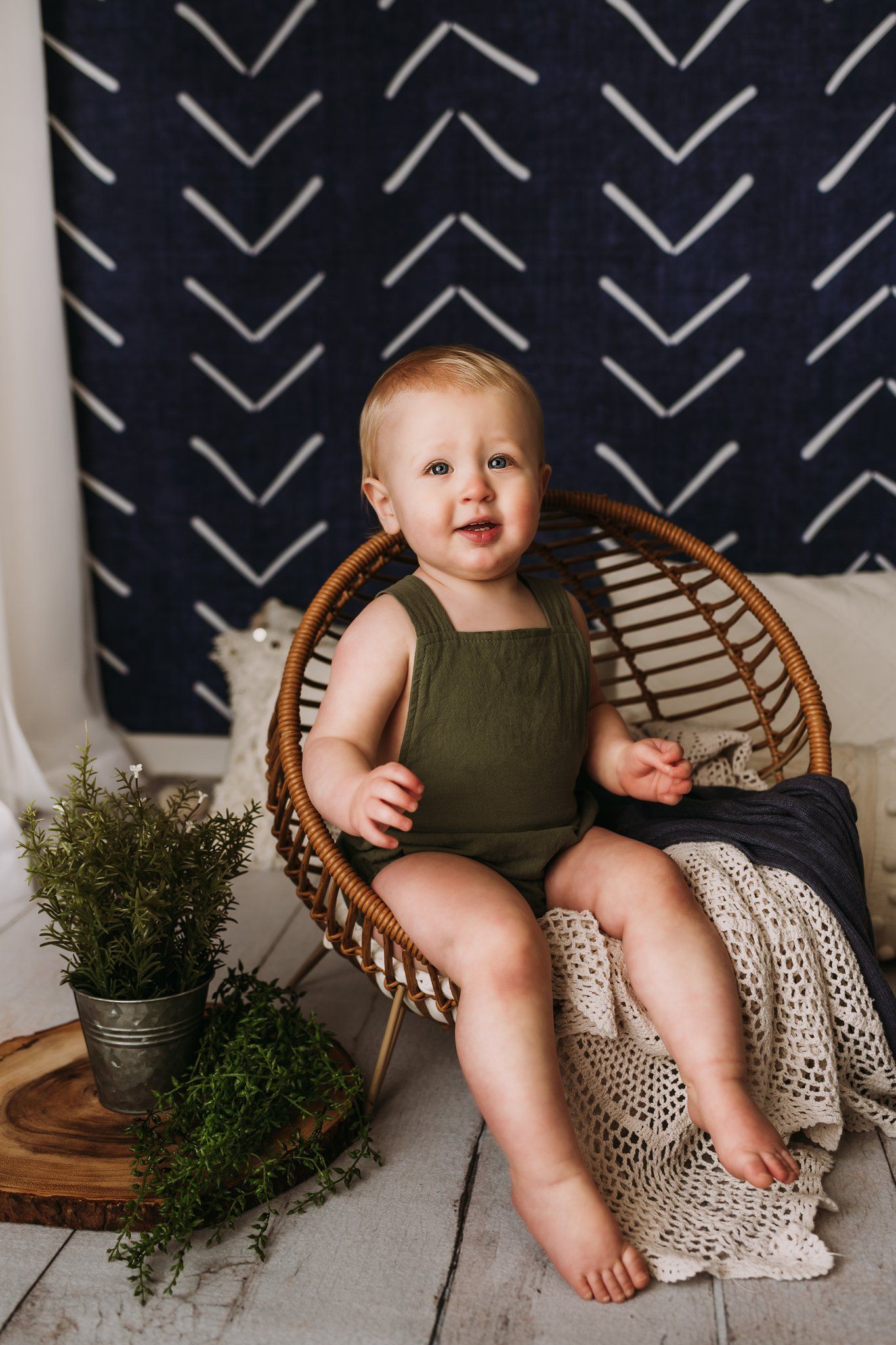 A toddler in green romper sits in a rattan chair with knit blankets and potted greenery.