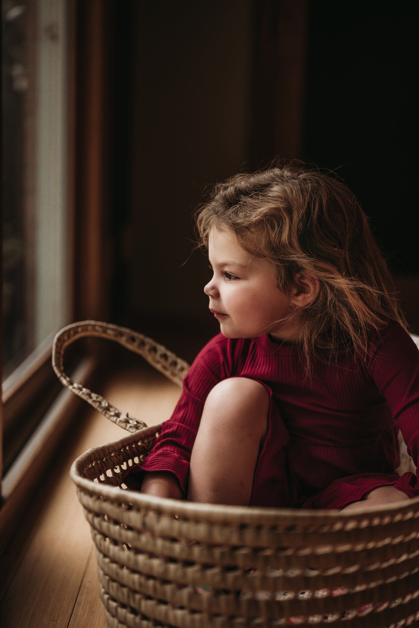 Child in red sweater sits thoughtfully in woven basket by window.