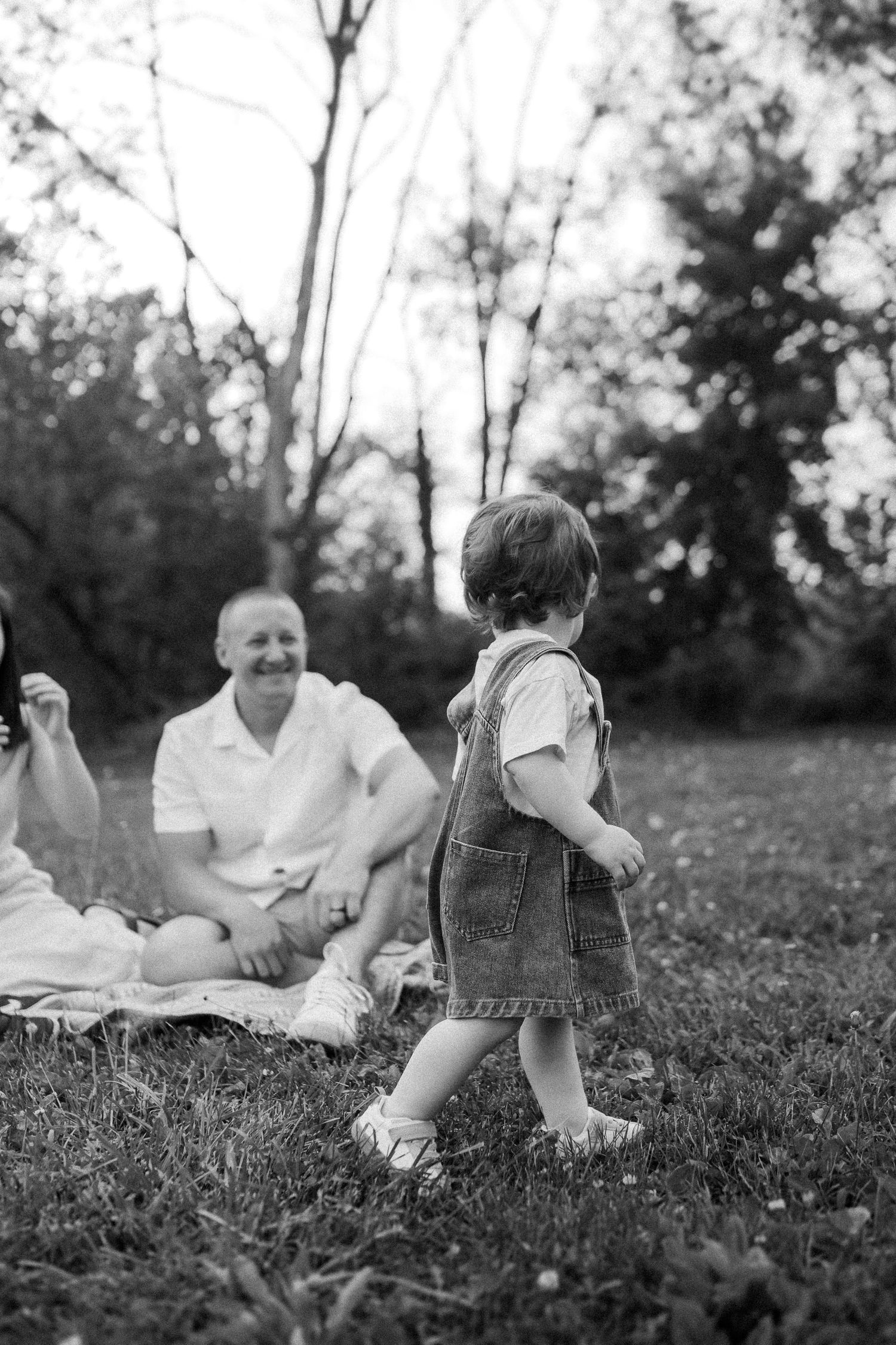A heartwarming black and white photo of a picnic scene in a grassy outdoor setting with a toddler in overalls.