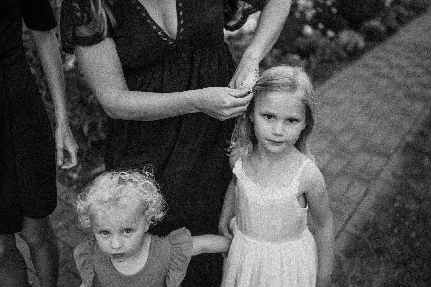 Black and white portrait of small children standing on brick pavers near flowering gardens.