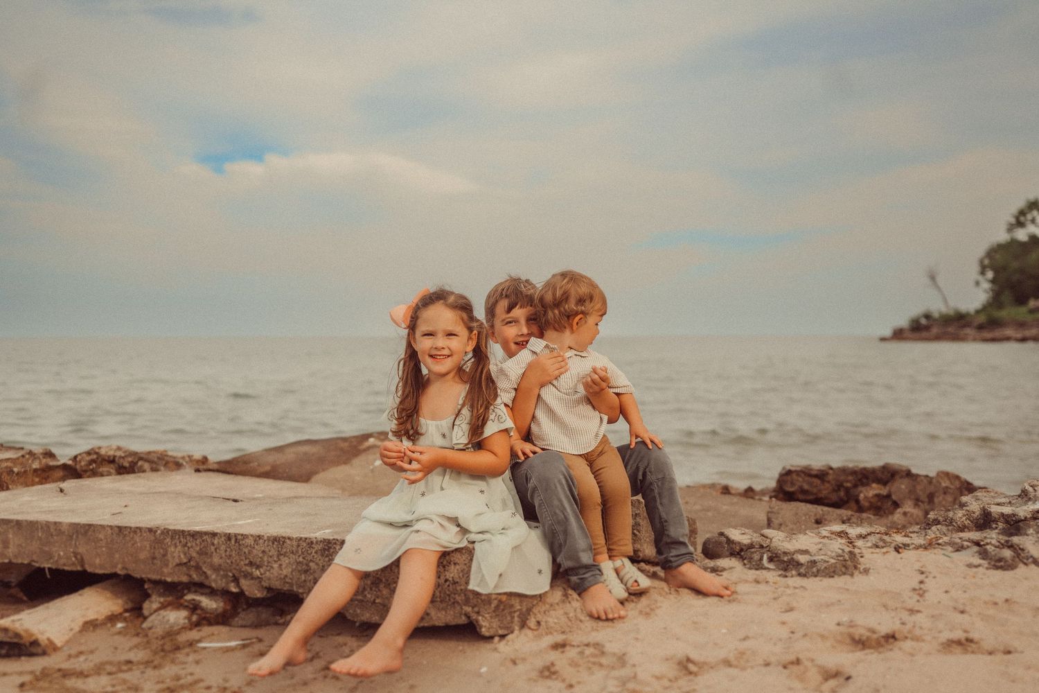 Three children sit together on rocky beach formations at sunset.