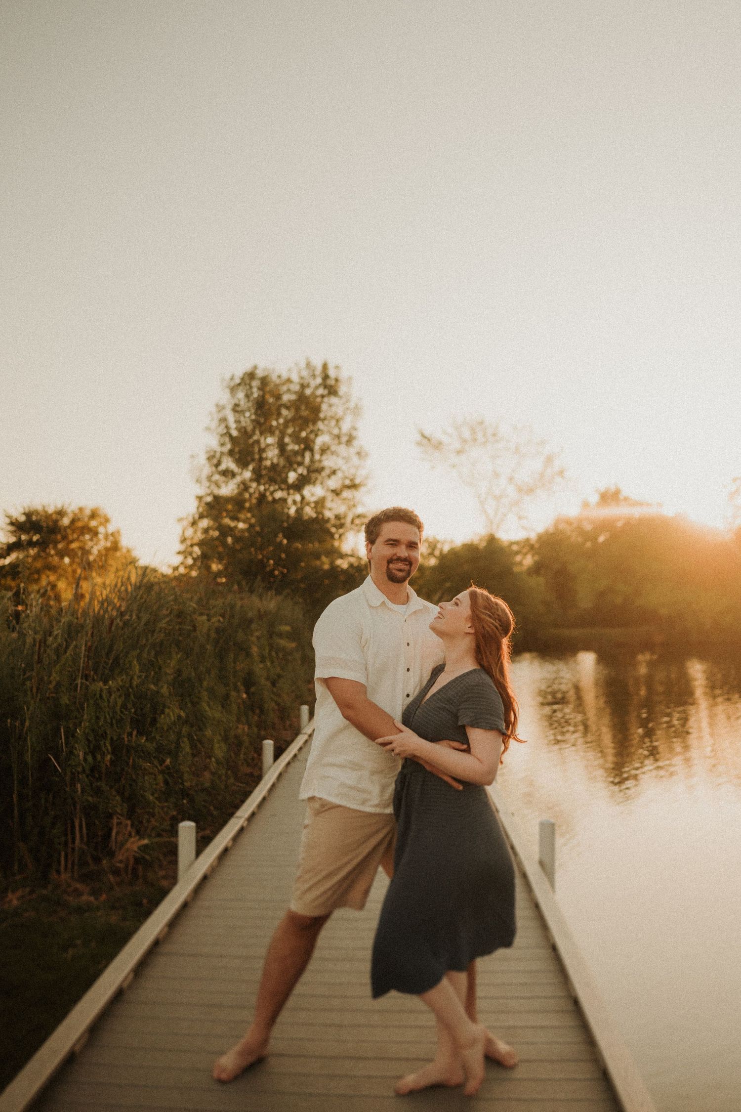A laughing couple dances barefoot on a wooden boardwalk at sunset.