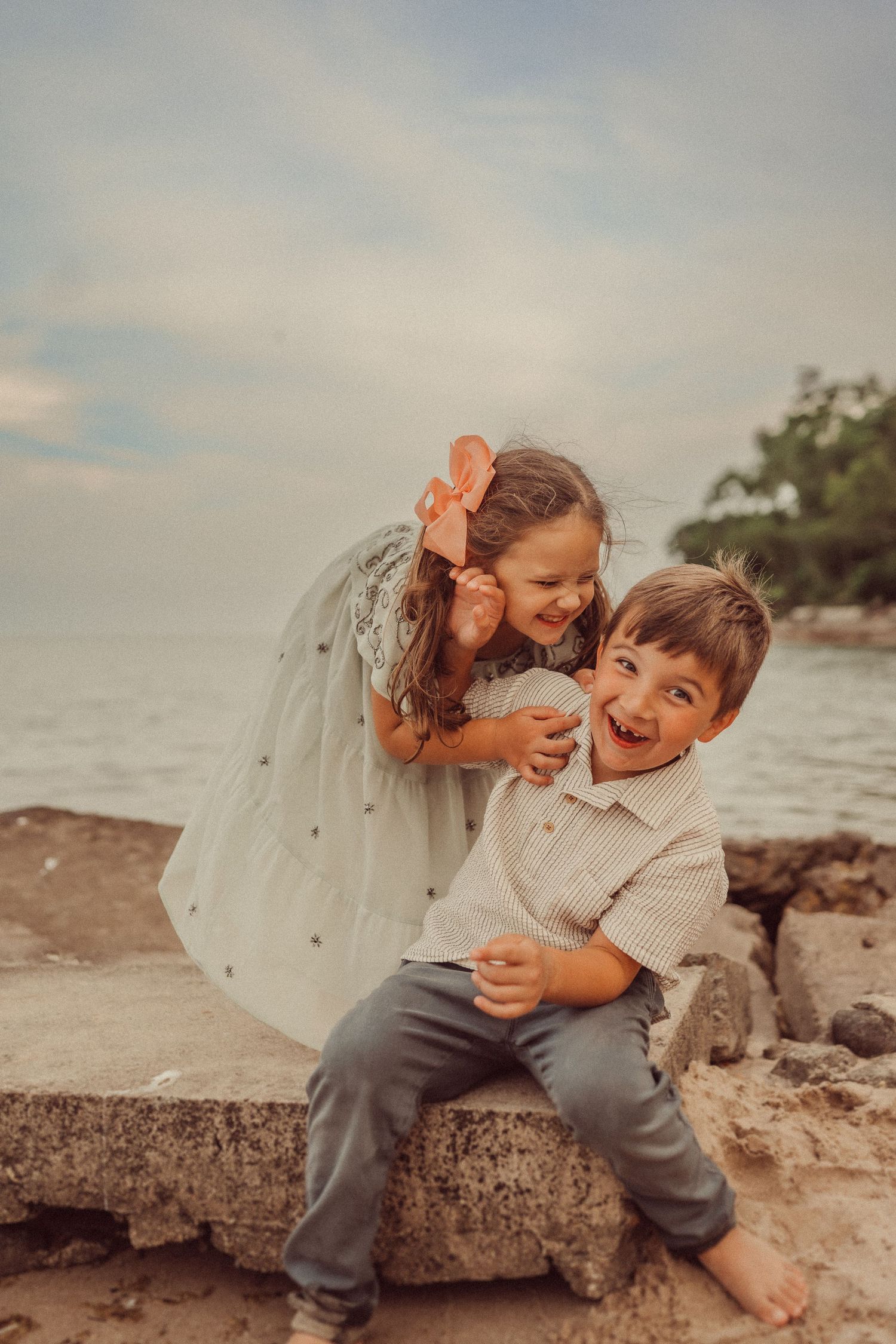 Two people share a playful moment laughing together while sitting on rocks by the ocean.