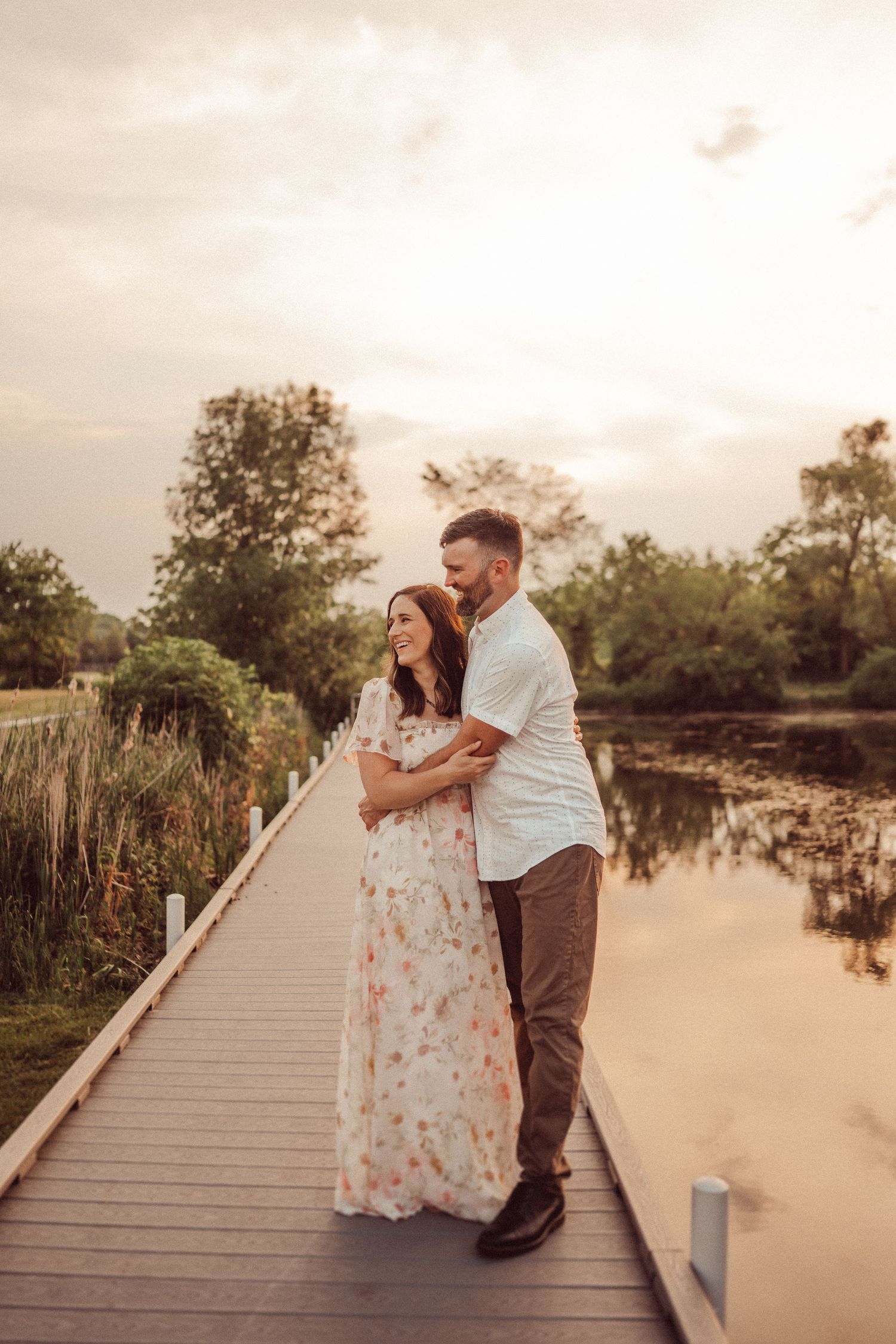 Romantic couple embracing on wooden boardwalk with calm water and sunset in background.
