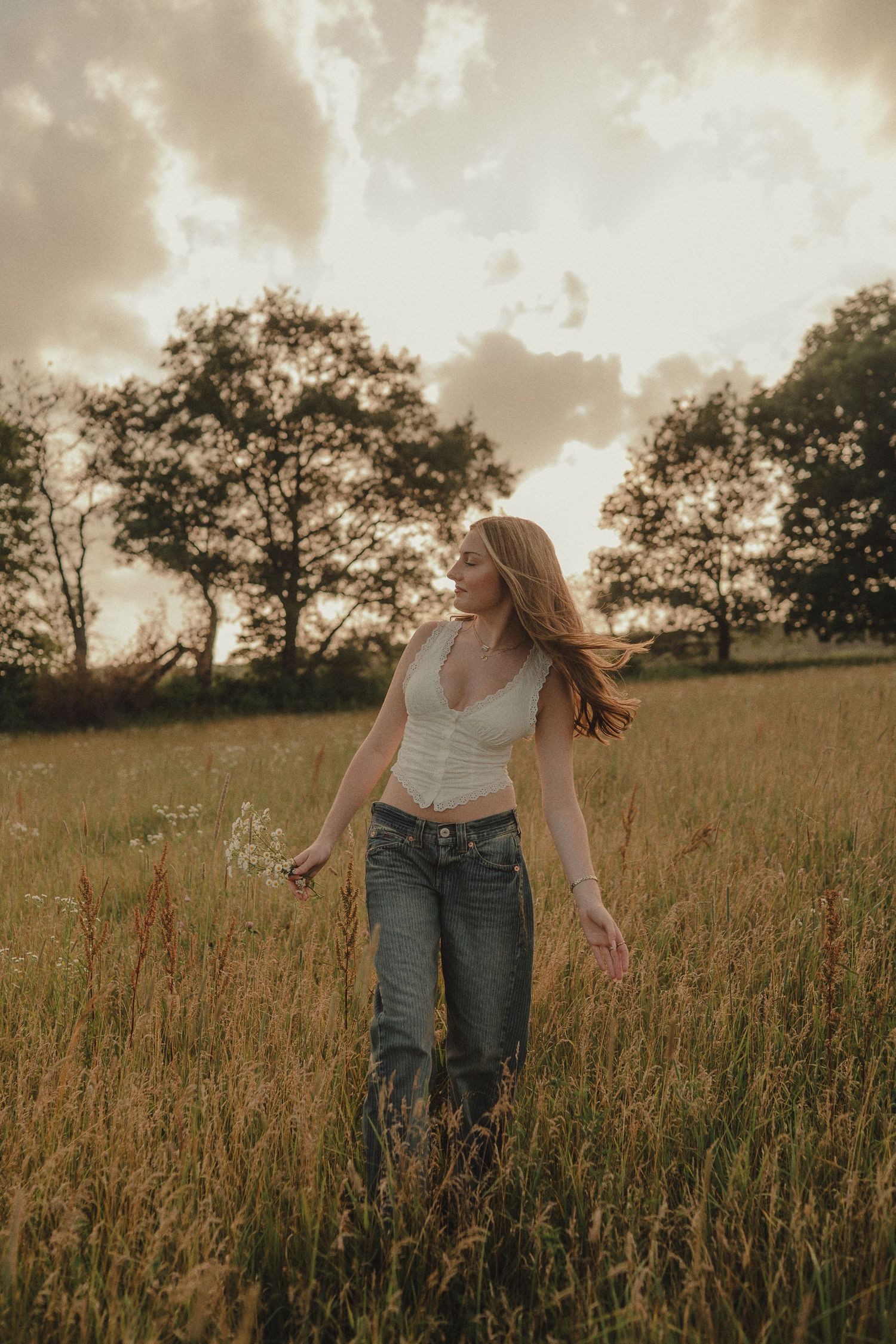 Young woman in white crop top and jeans walking through golden meadow at sunset with wind in hair.