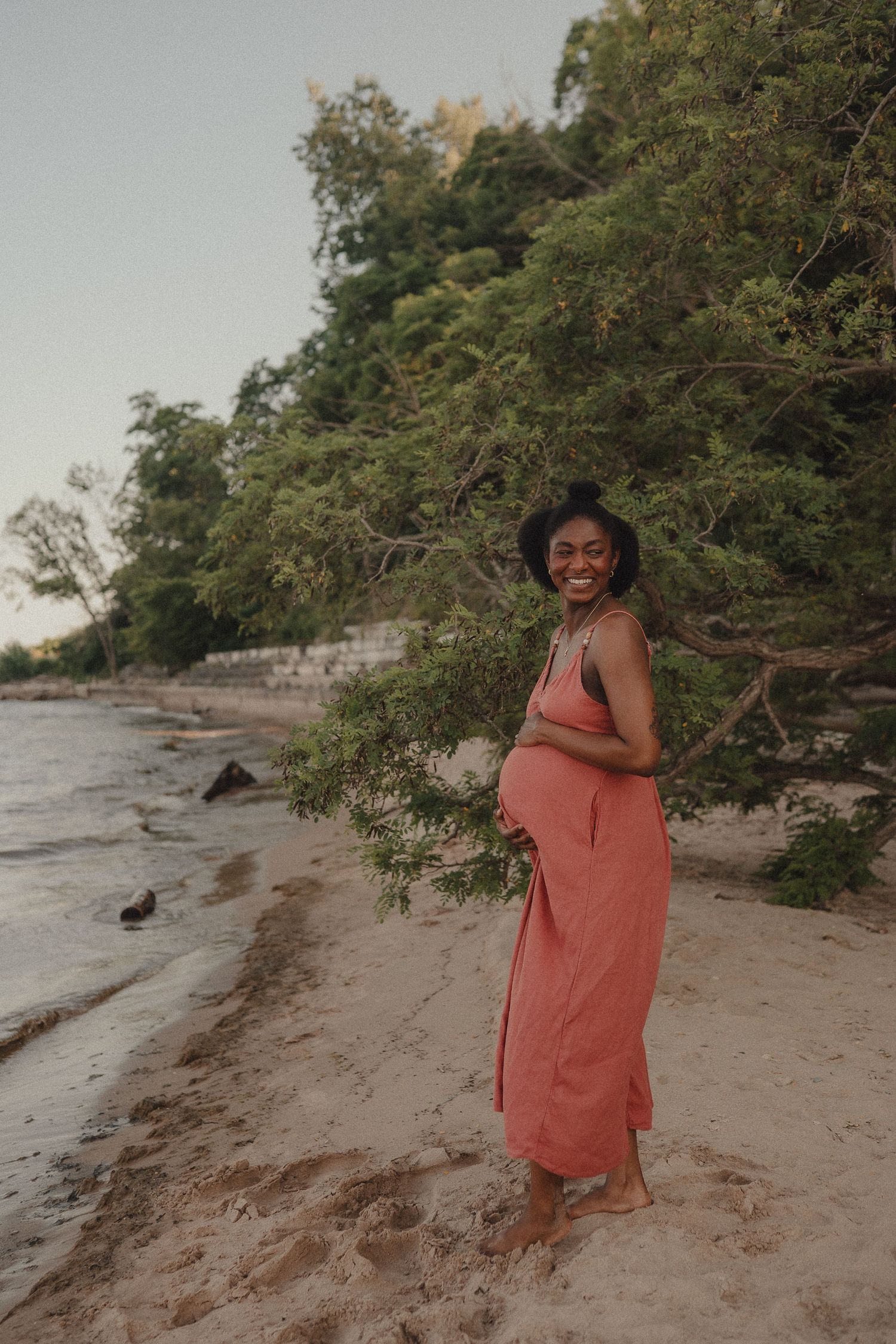 Smiling pregnant woman in coral maxi dress standing barefoot on sandy beach with lush green trees behind her.