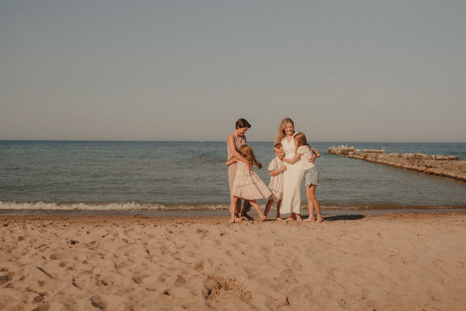 Family gathering on a sandy beach near calm ocean waters during golden hour, dressed in light summer outfits.