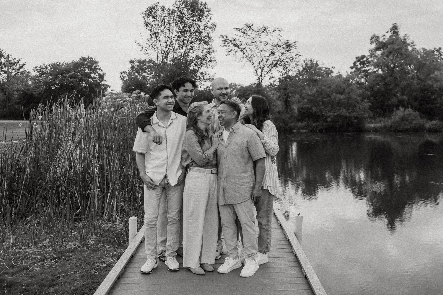 Black and white photo of a family posing together on a wooden dock by a pond surrounded by lush trees.
