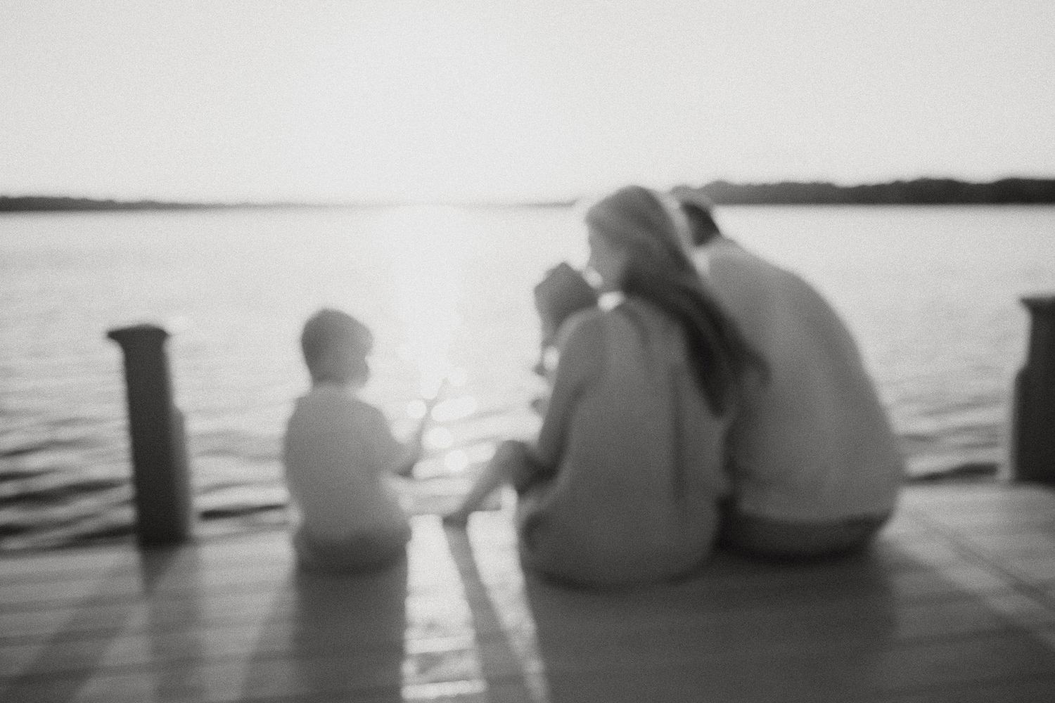 Family sits together on a dock watching a serene lake sunset in a soft-focus black and white photo.