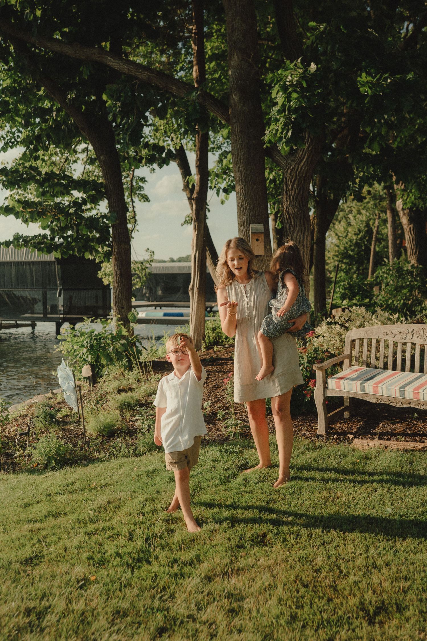 Mother and two children playing barefoot in lush green backyard near lakeside garden.