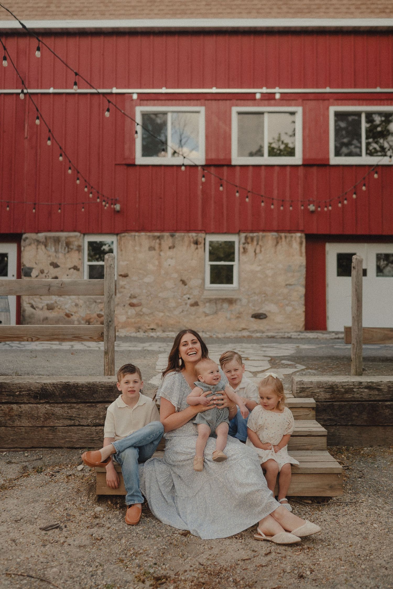 Smiling mother in floral dress sitting on wooden steps with four children in front of a red barn with string lights.