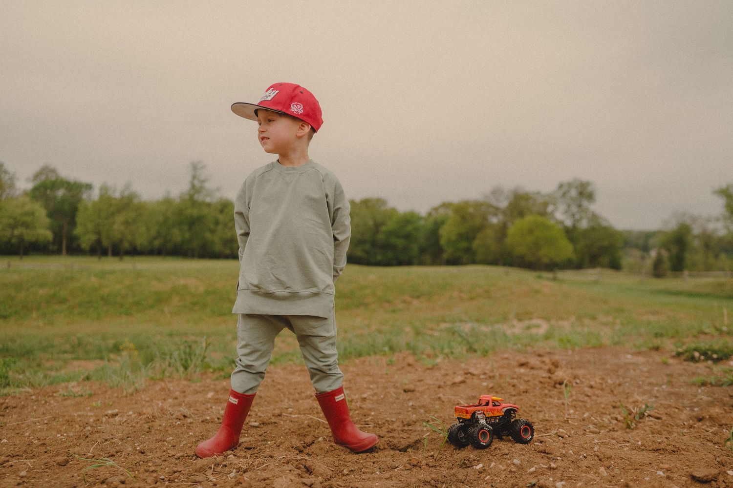 Young boy in red cap and boots stands in muddy field with toy monster truck on overcast day.