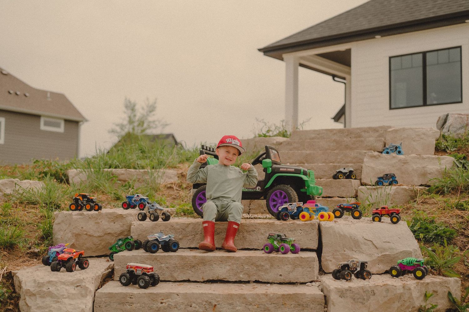 Young boy in red boots stands among colorful monster truck toy collection on stone steps.