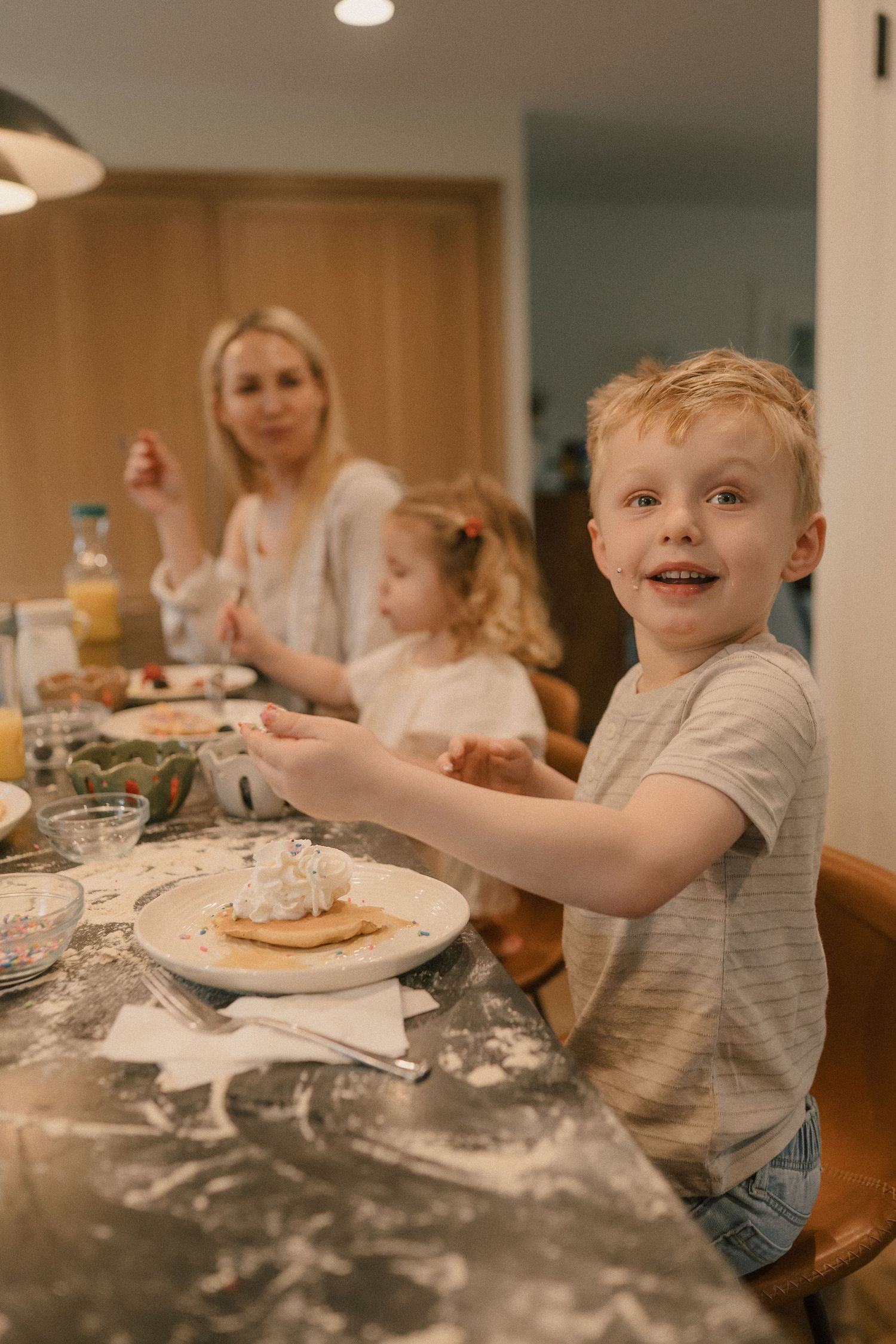 Smiling young boy reaching for pancakes at a family breakfast table with mother and sister behind him.