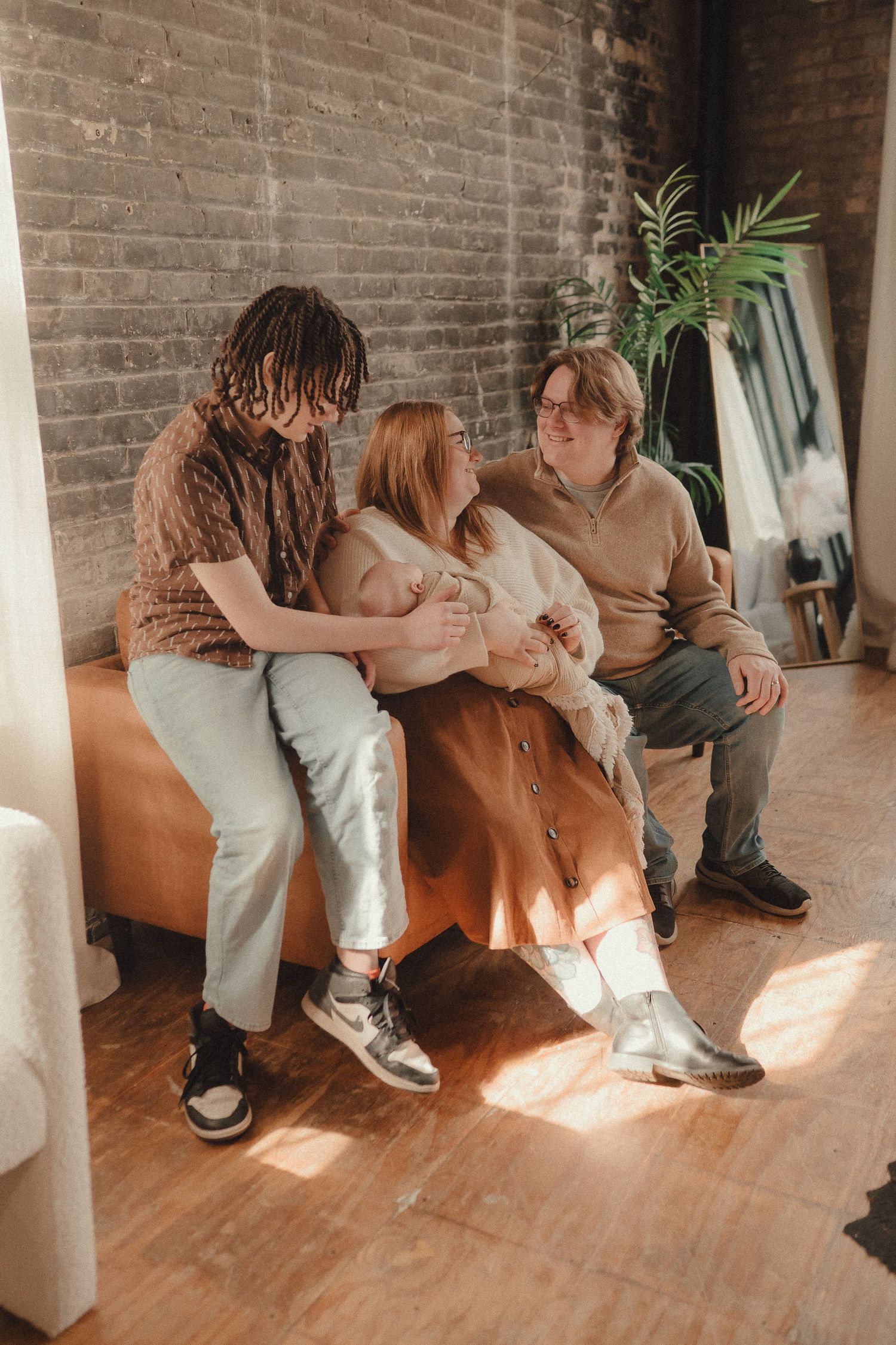 A family of three sits together on a couch, smiling and holding a newborn baby in a warm sunlit room.