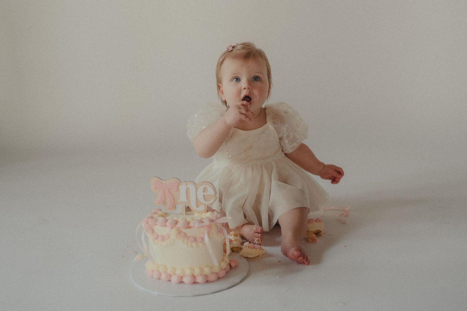 Cute baby girl in a white tulle dress tastes frosting from her pink smash cake during first birthday photos.