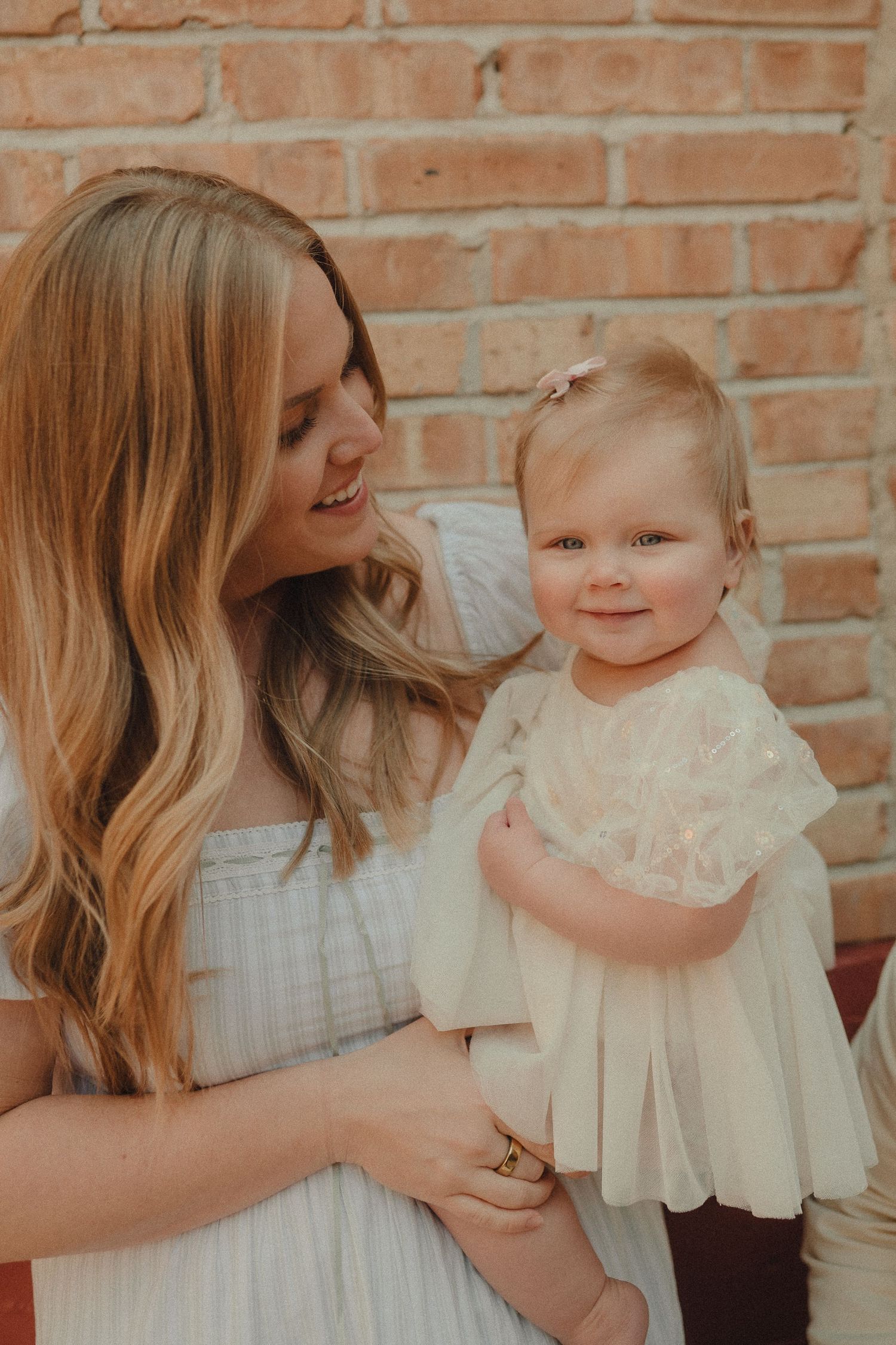 A smiling mother with long blonde hair holds her baby girl in a cream lace dress against a brick wall.