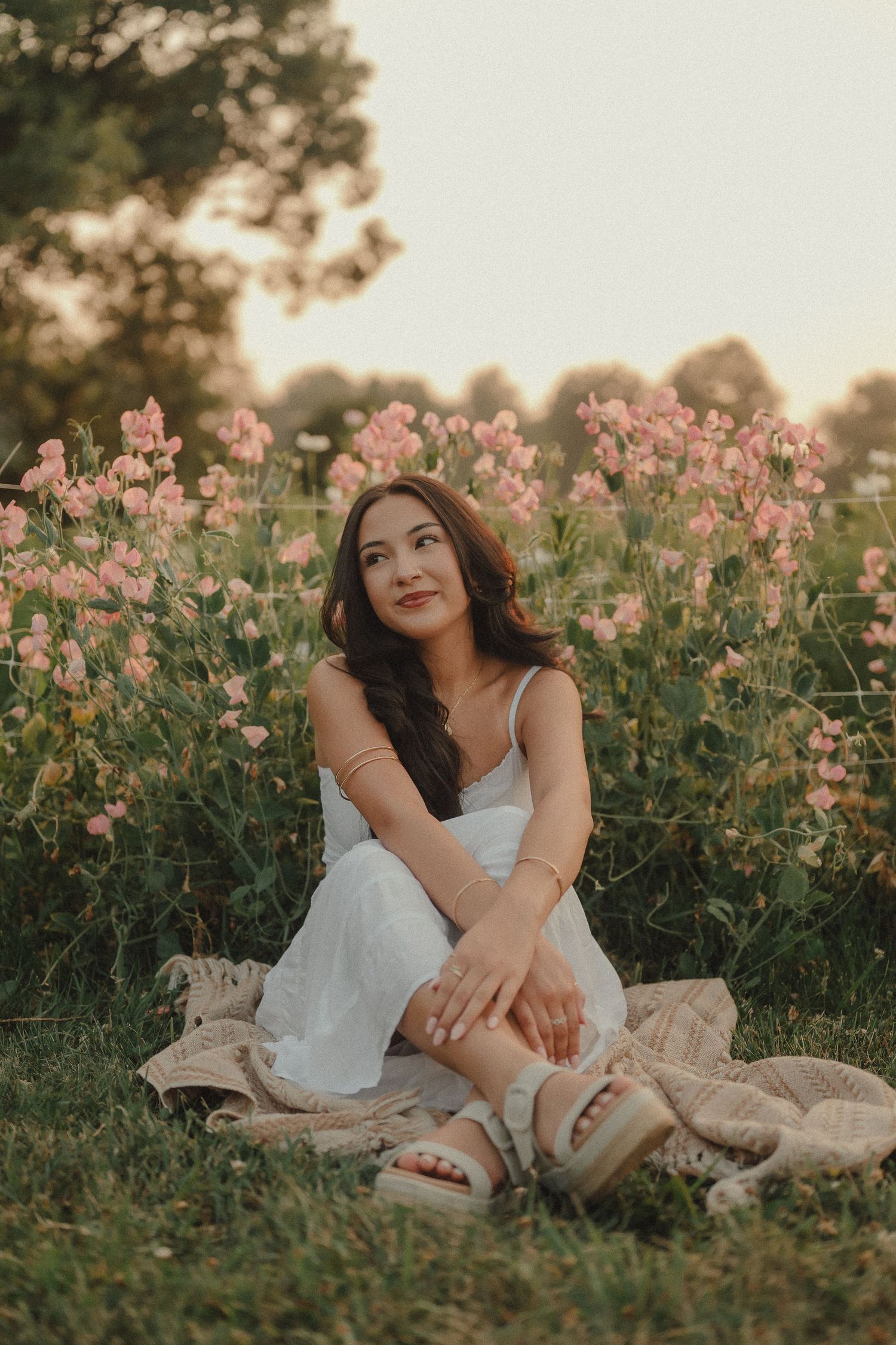 Young woman in white dress sitting on blanket among pink wildflowers at golden hour.
