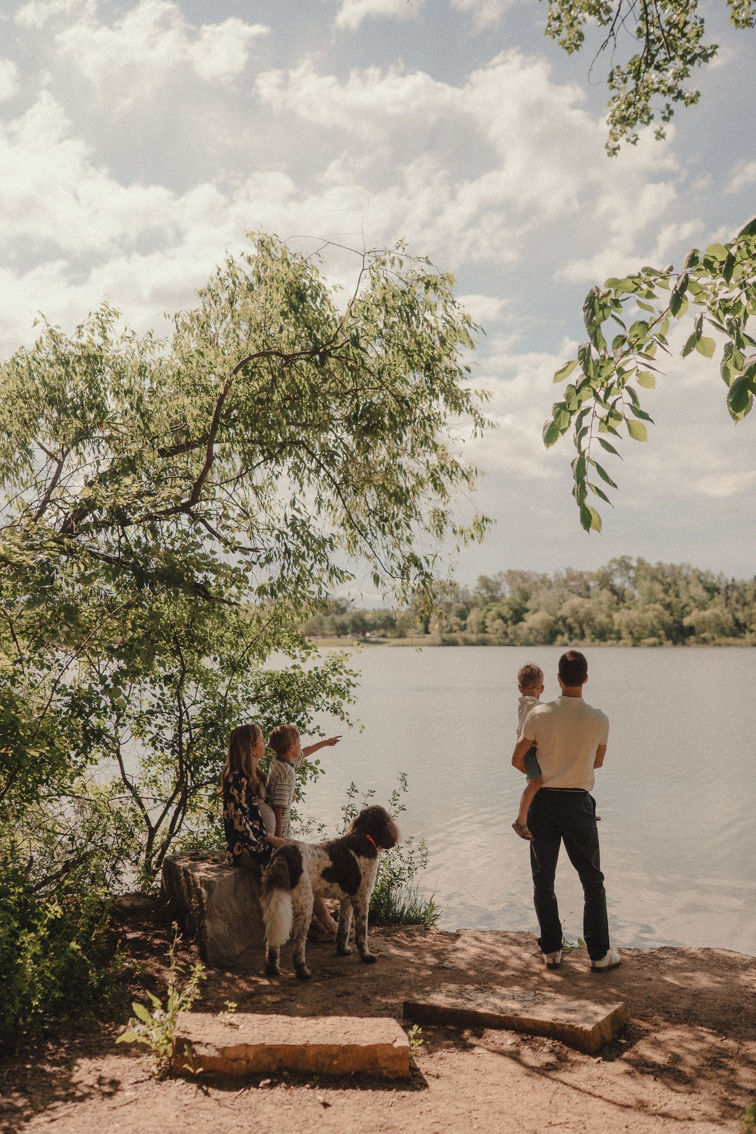 A father holds a toddler while a woman and dog explore the rocky shore of a tree-lined lake.