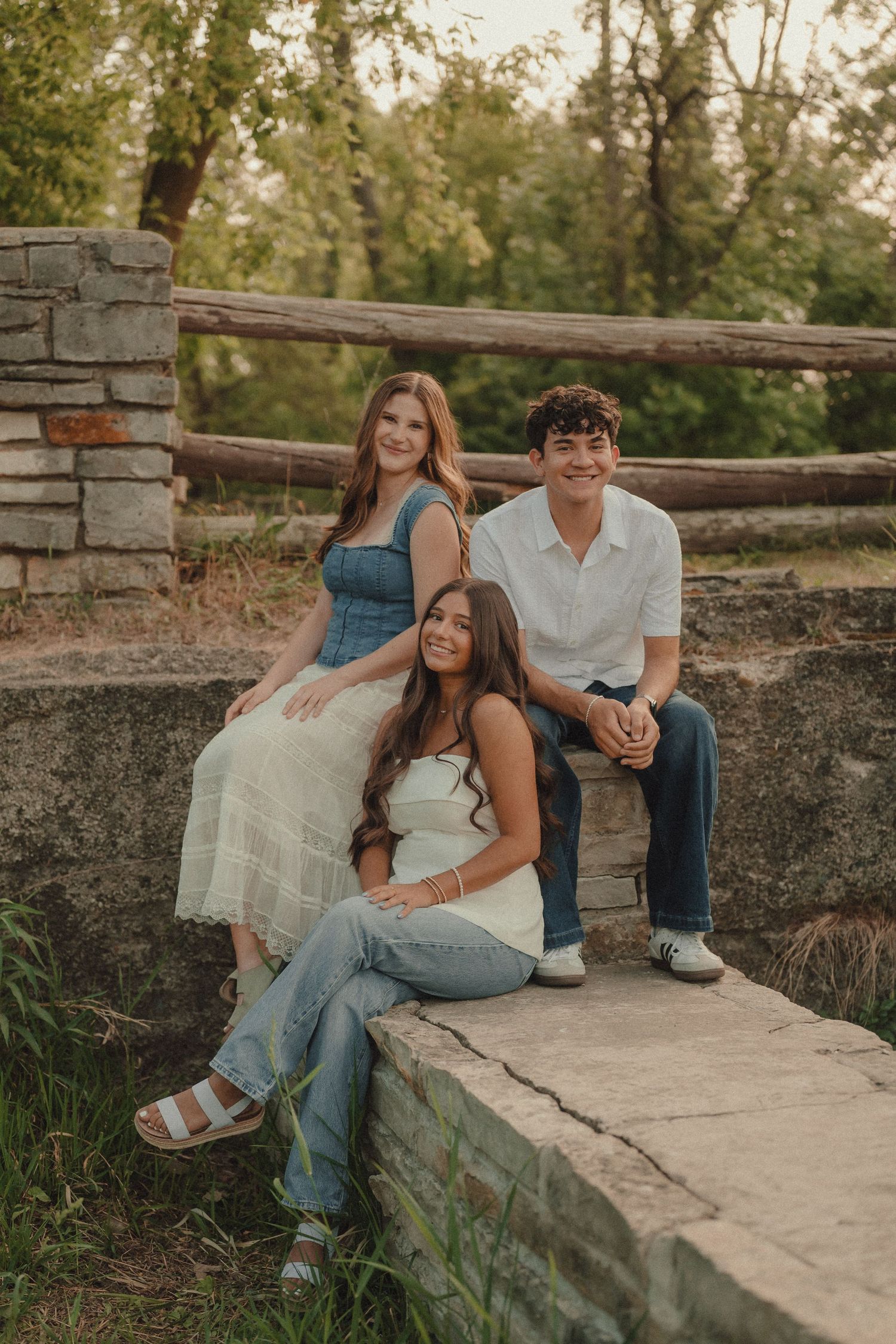 Three smiling teens pose on a stone bridge outdoors, two girls and a boy in casual summer outfits.