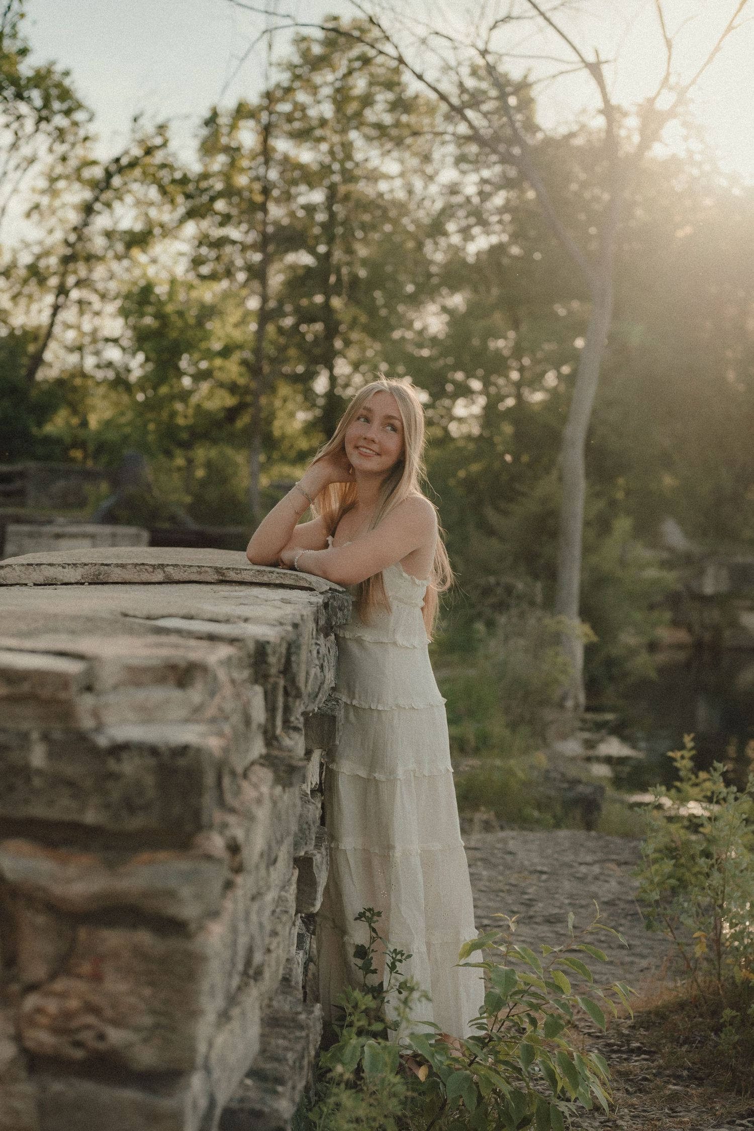 Young blonde woman in white maxi dress leaning on stone wall surrounded by lush green trees in golden hour sunlight.