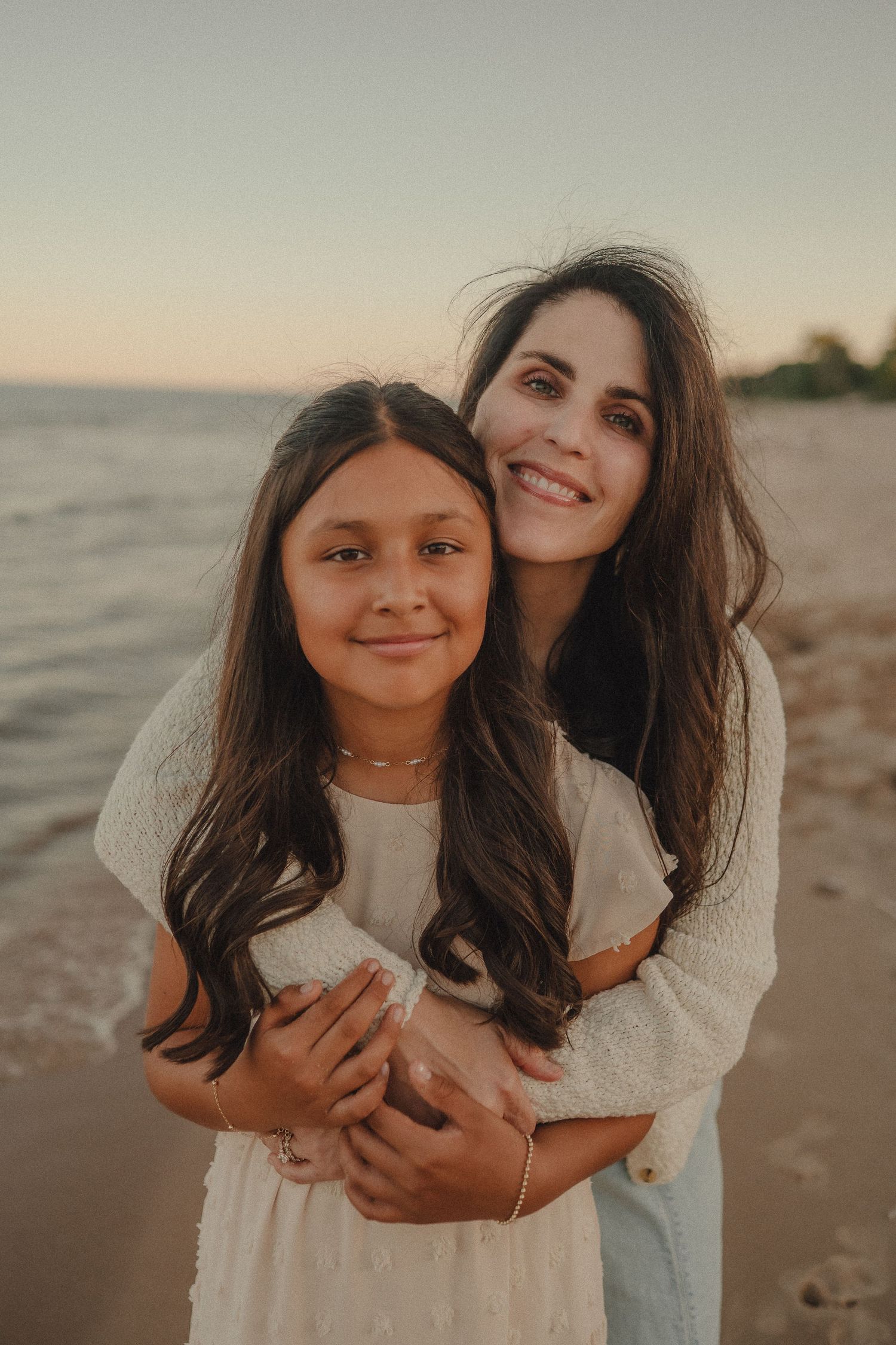 Smiling mother hugging young daughter from behind on beach at sunset in matching white dresses.