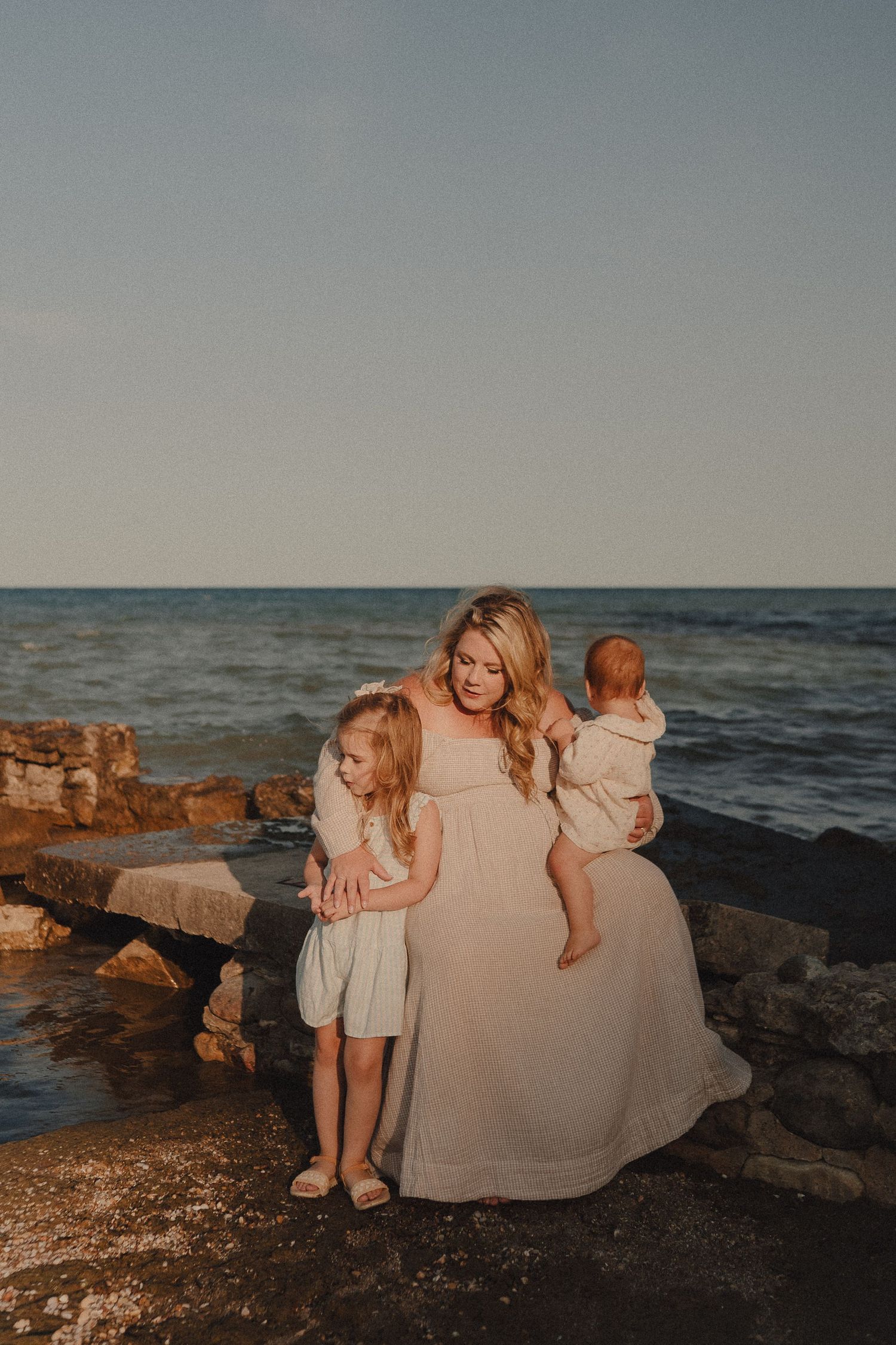 A blonde mother in a white dress holds her baby while her young daughter hugs her on rocky beach at sunset.
