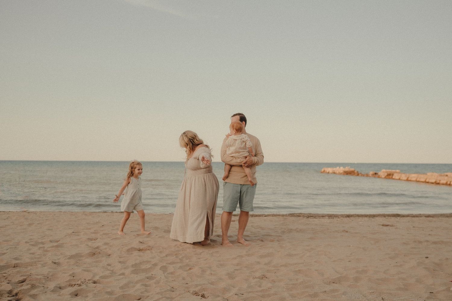 Family of four standing on sandy beach at sunset, parents with baby and young daughter by the sea.