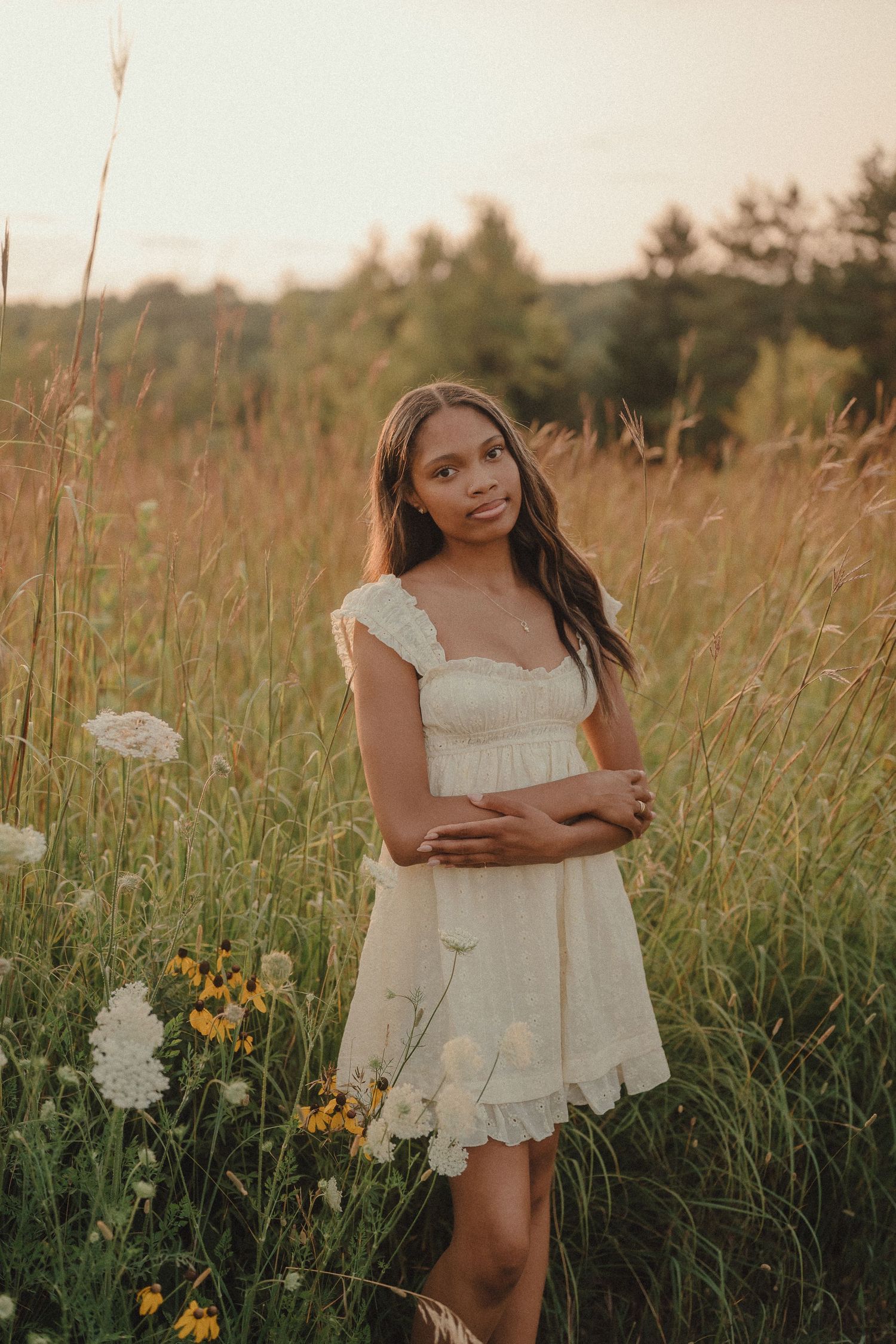 Young woman in white lace mini dress standing among wildflowers in golden meadow at sunset.
