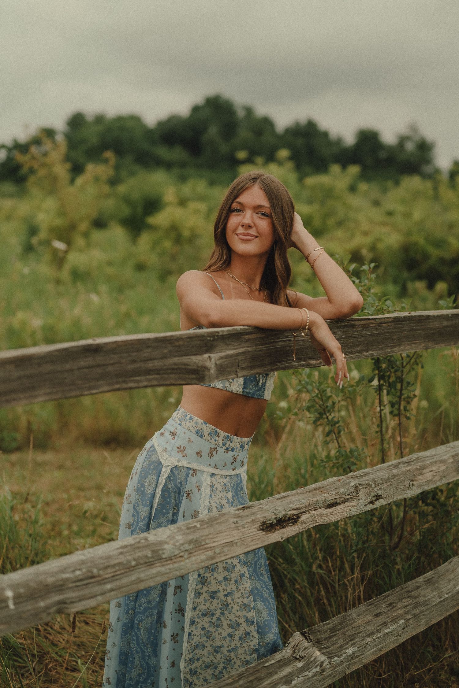 Young woman in blue floral outfit leaning on rustic wooden fence in lush green meadow.