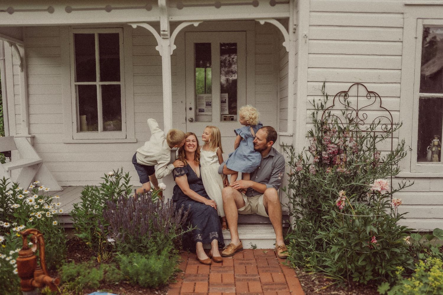 Family portrait on vintage farmhouse porch with brick walkway and flowering garden beds.