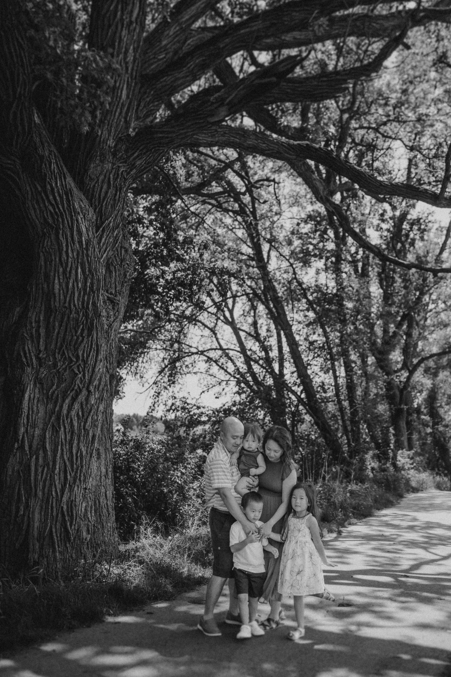 Black and white portrait of a family standing beneath a large tree on a nature path.