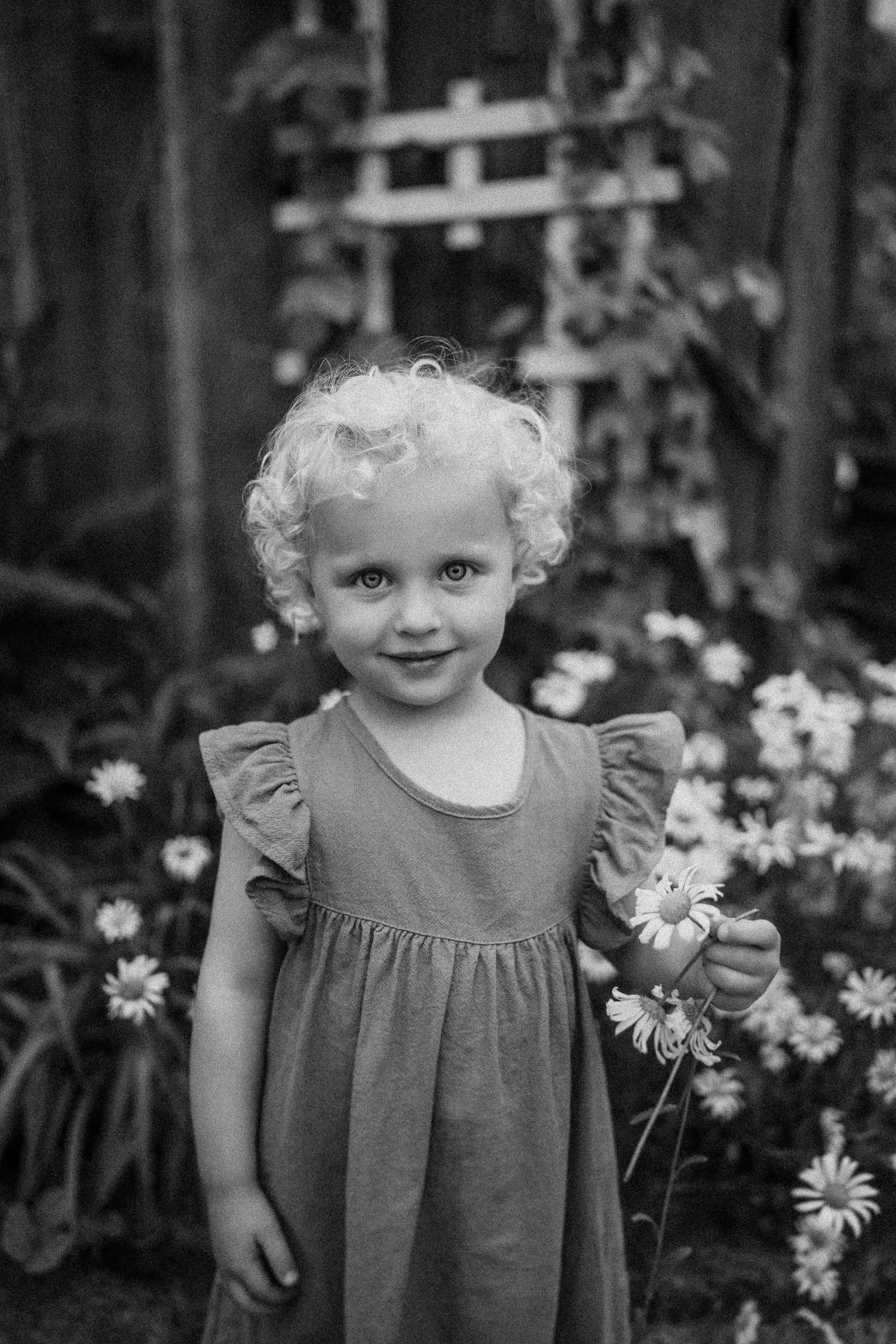 Black and white portrait of a child in a dress standing by blooming daisies and a garden trellis.