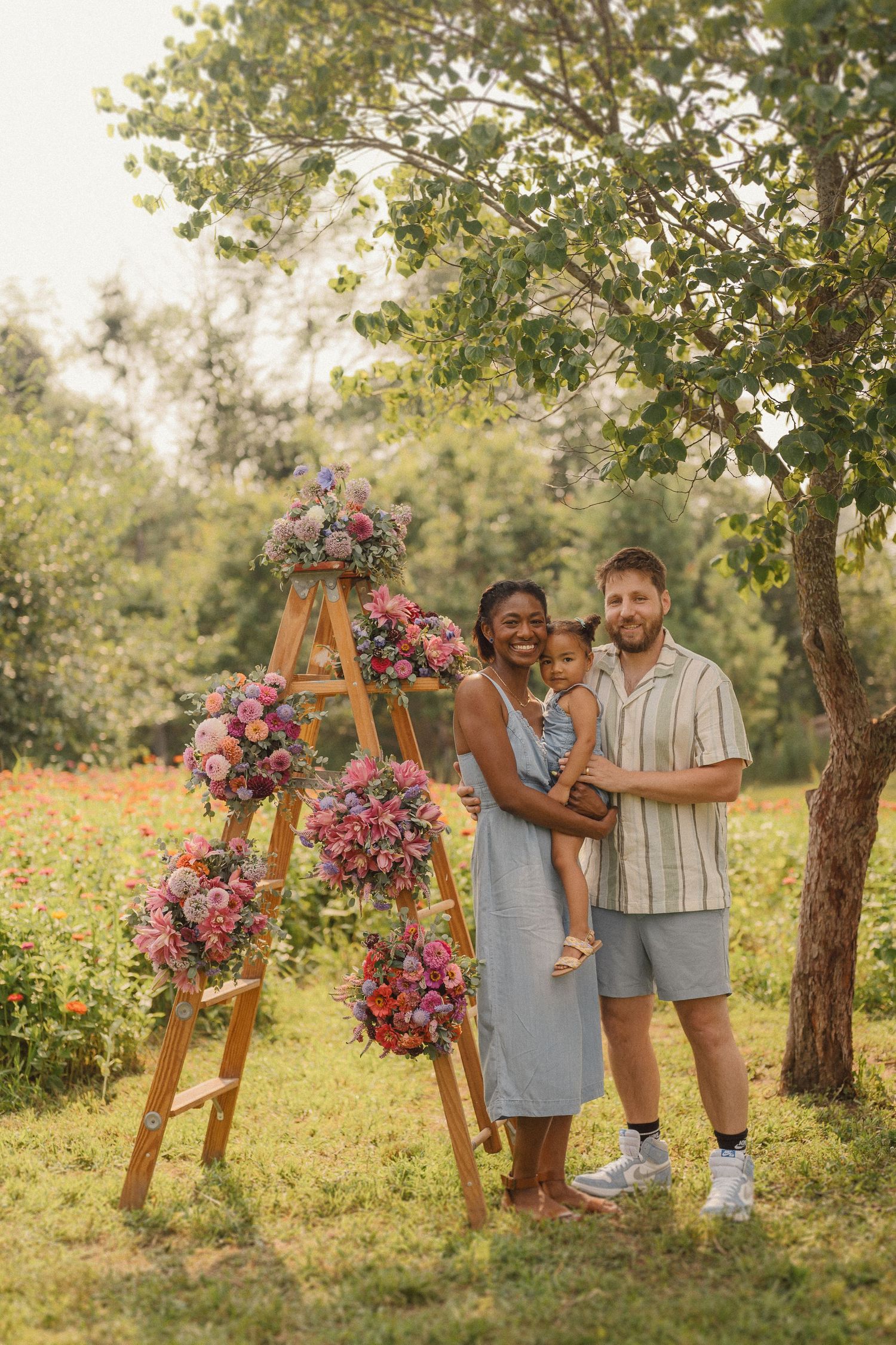 Happy couple poses beside wooden easel adorned with pink floral arrangement in sunlit garden setting.
